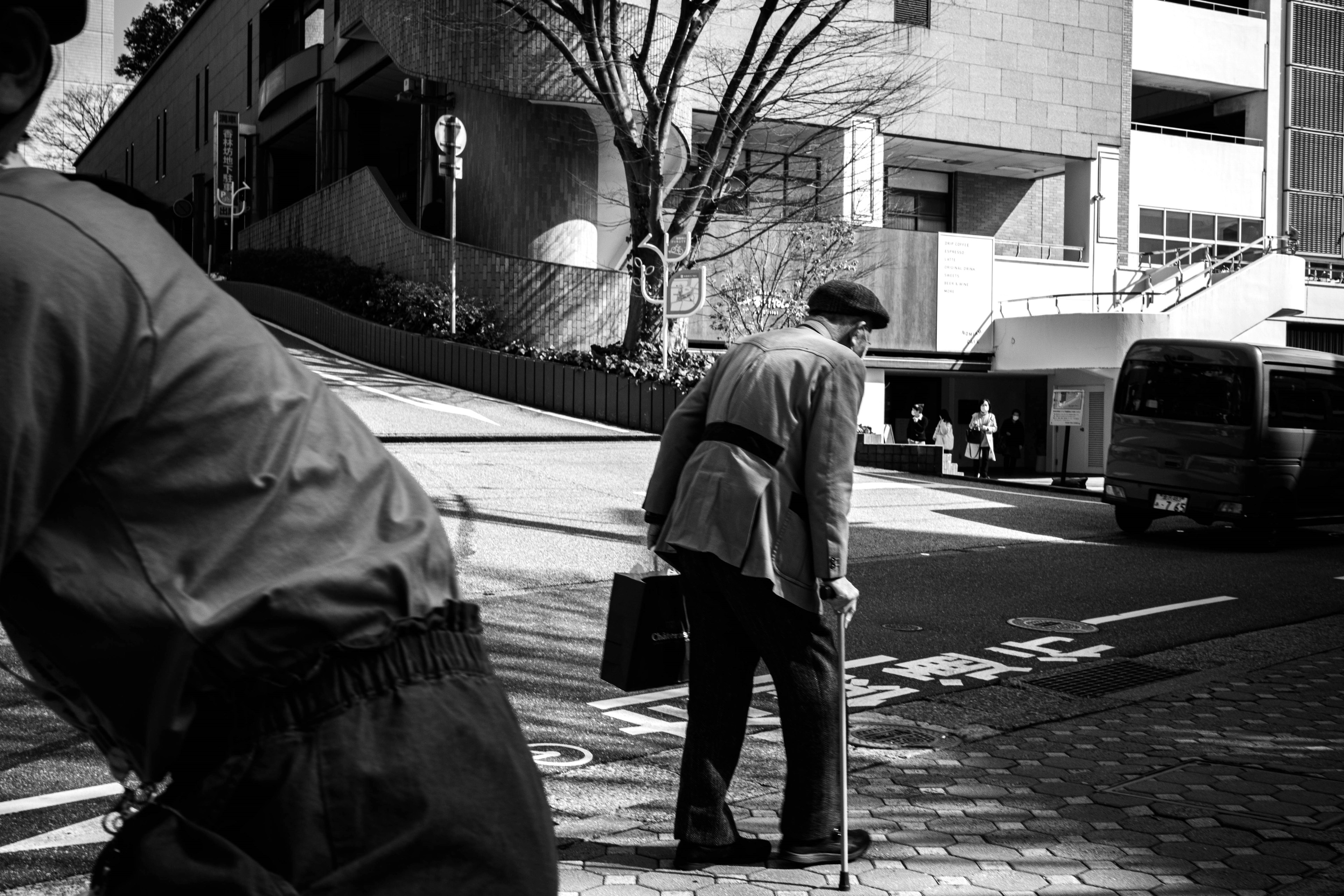 Black and white image of an elderly man walking with a cane in an urban setting with buildings and a tree