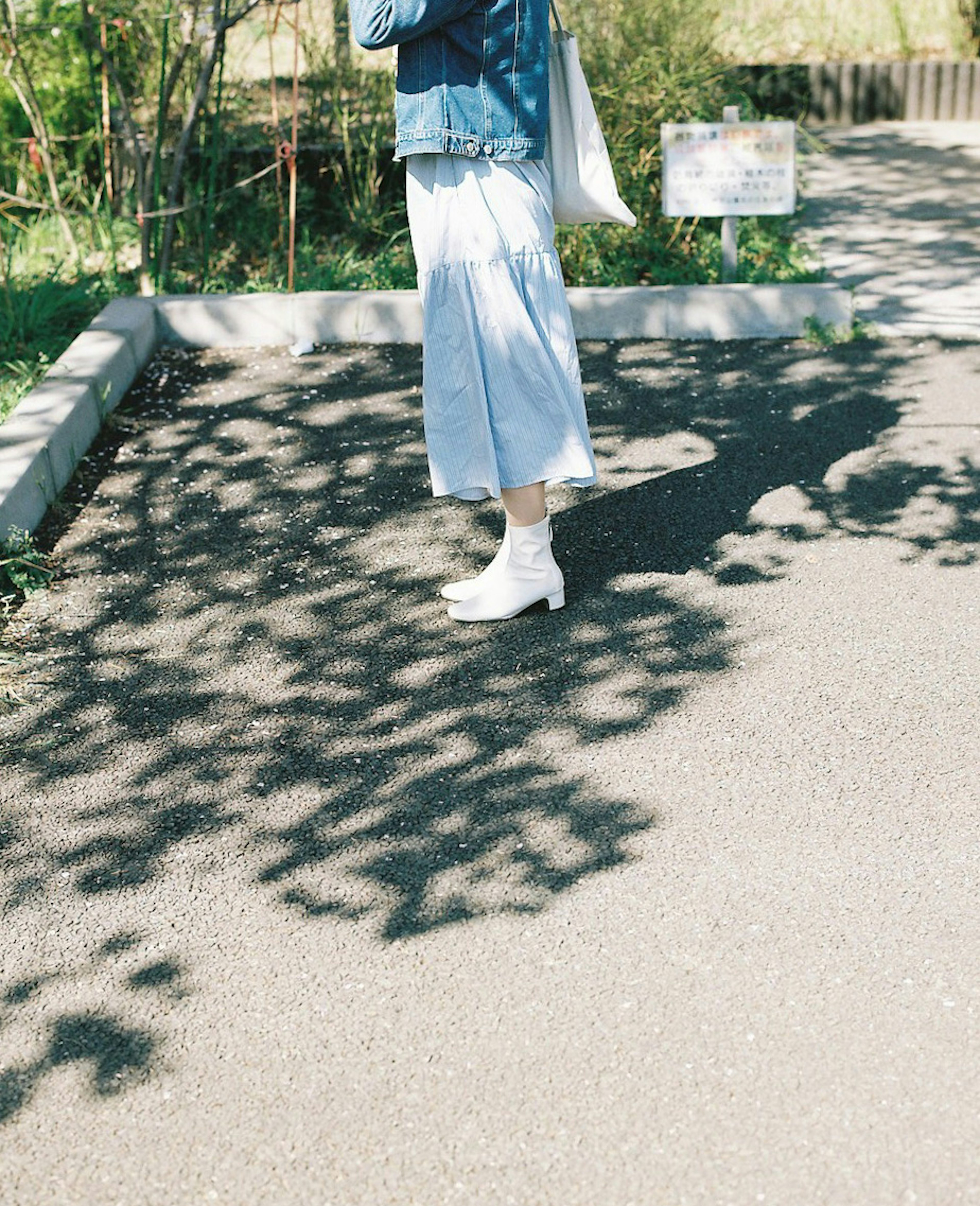 A woman wearing a denim jacket and white shoes stands on a path with shadows