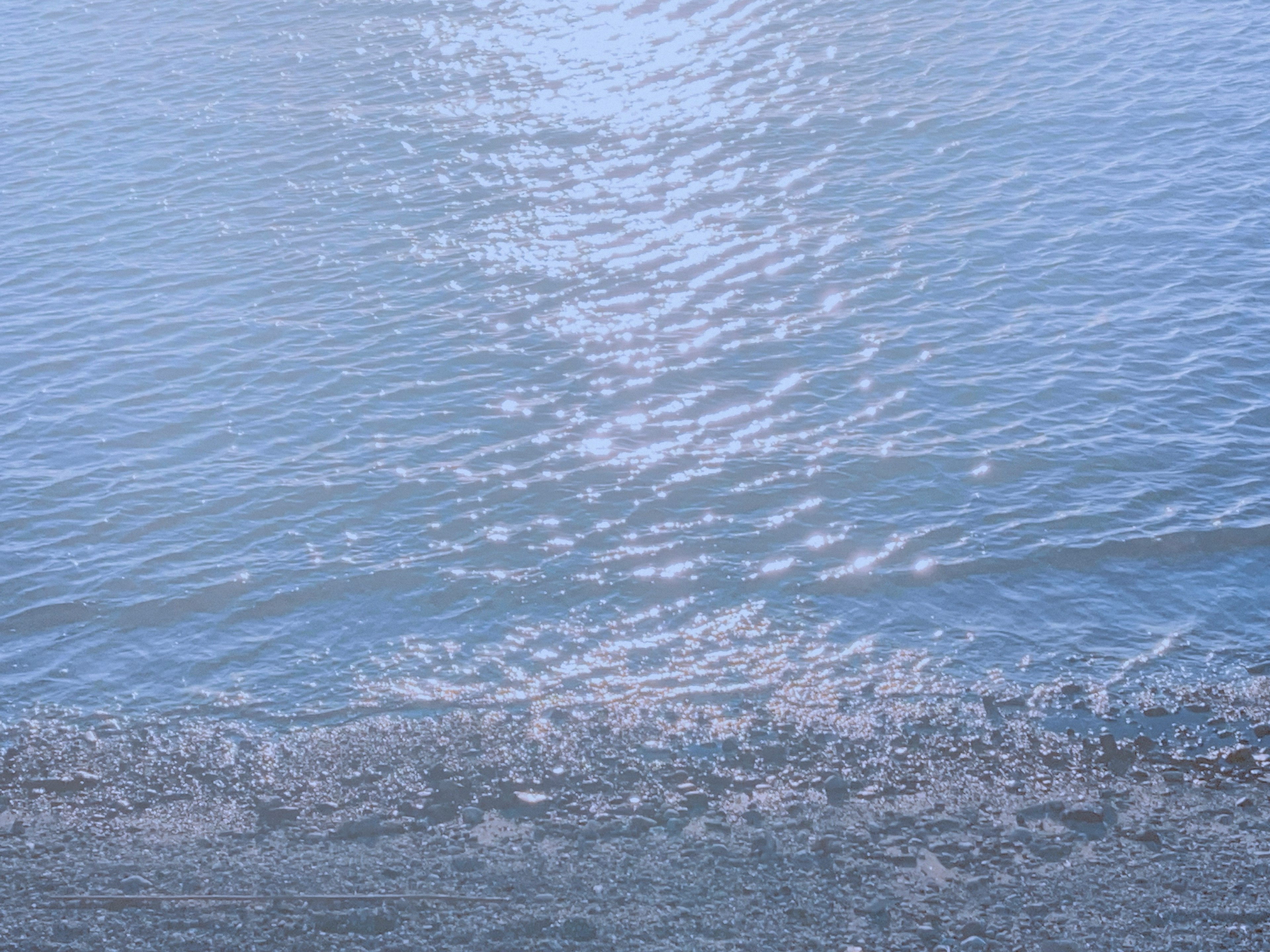 Blaue Wasseroberfläche mit Lichtreflexion und sandigem Ufer