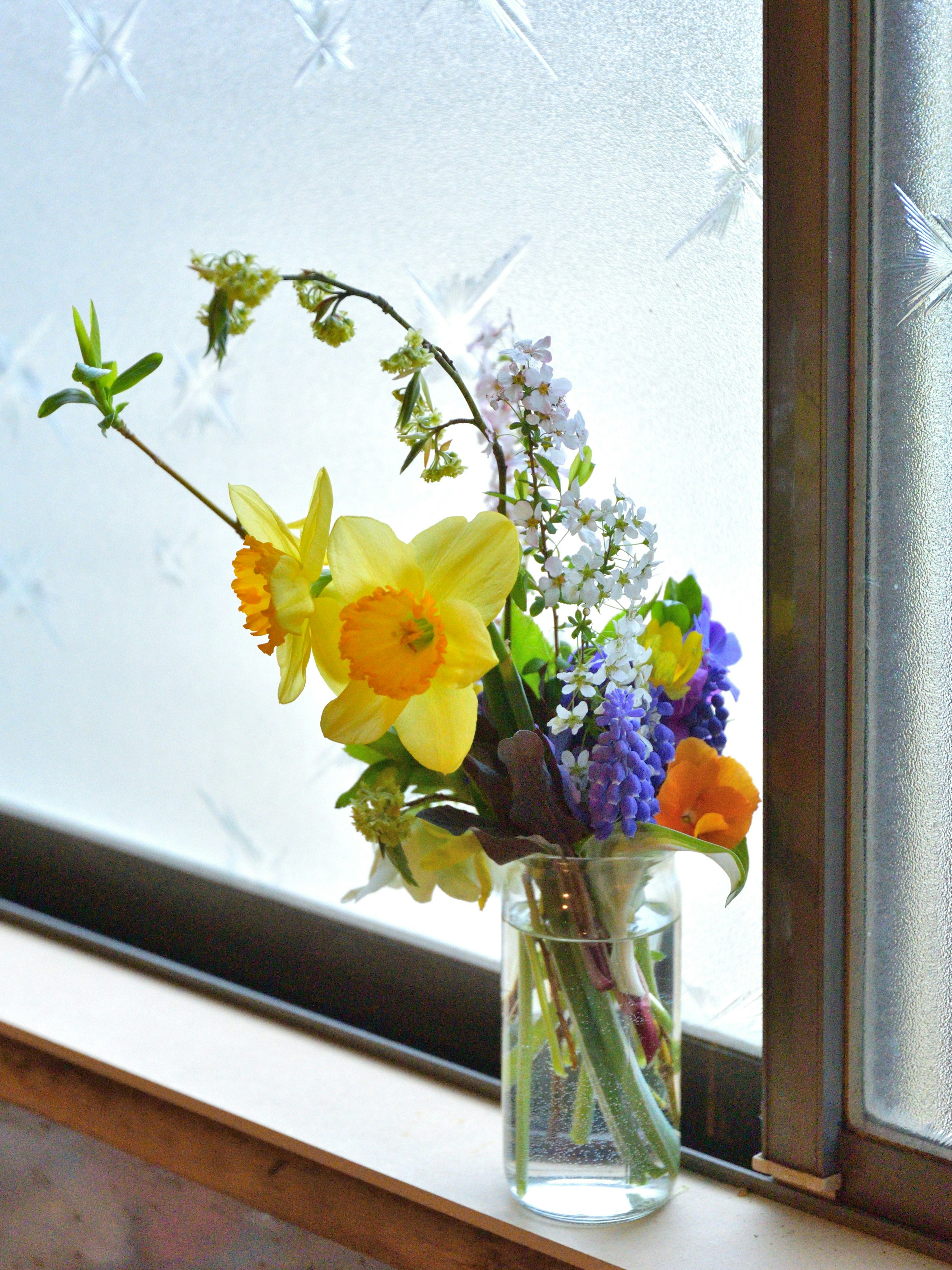 A bouquet of yellow daffodils and various colorful flowers in a glass vase by the window