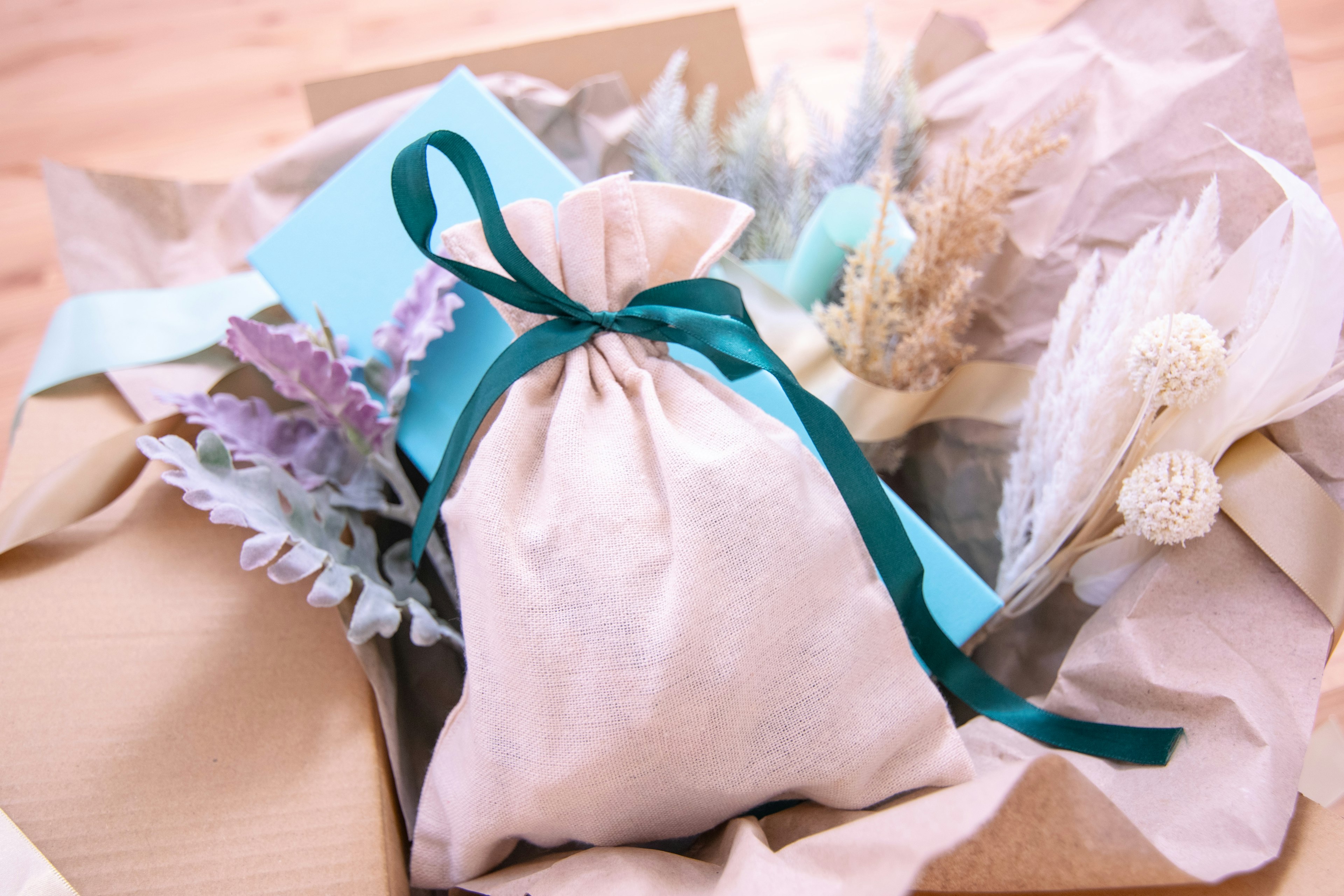 A gift box containing a white fabric pouch tied with a blue ribbon surrounded by dried flowers and decorative packaging