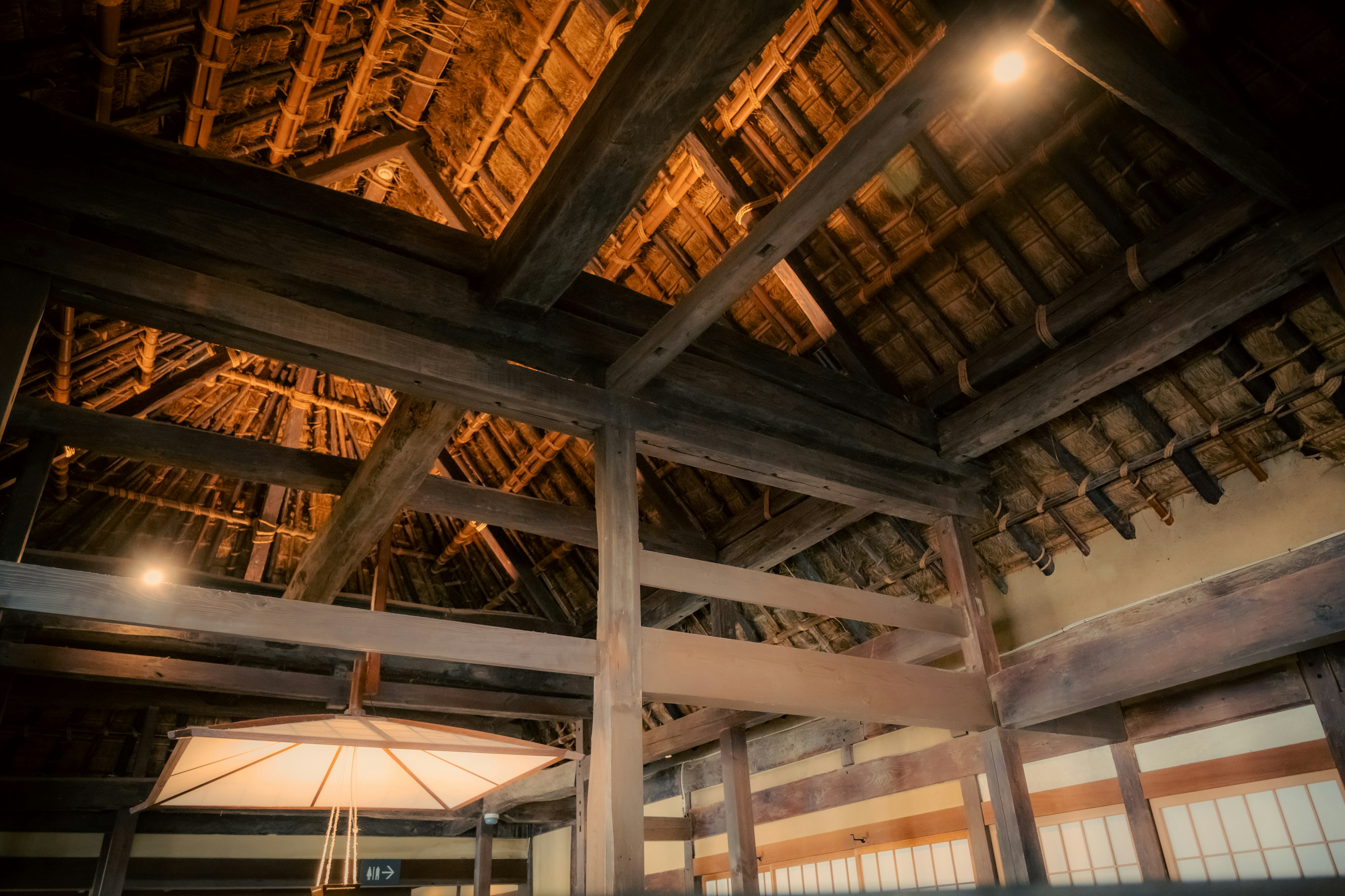 Vista interior de una estructura tradicional japonesa con techo de paja que muestra vigas de madera y detalles de paja