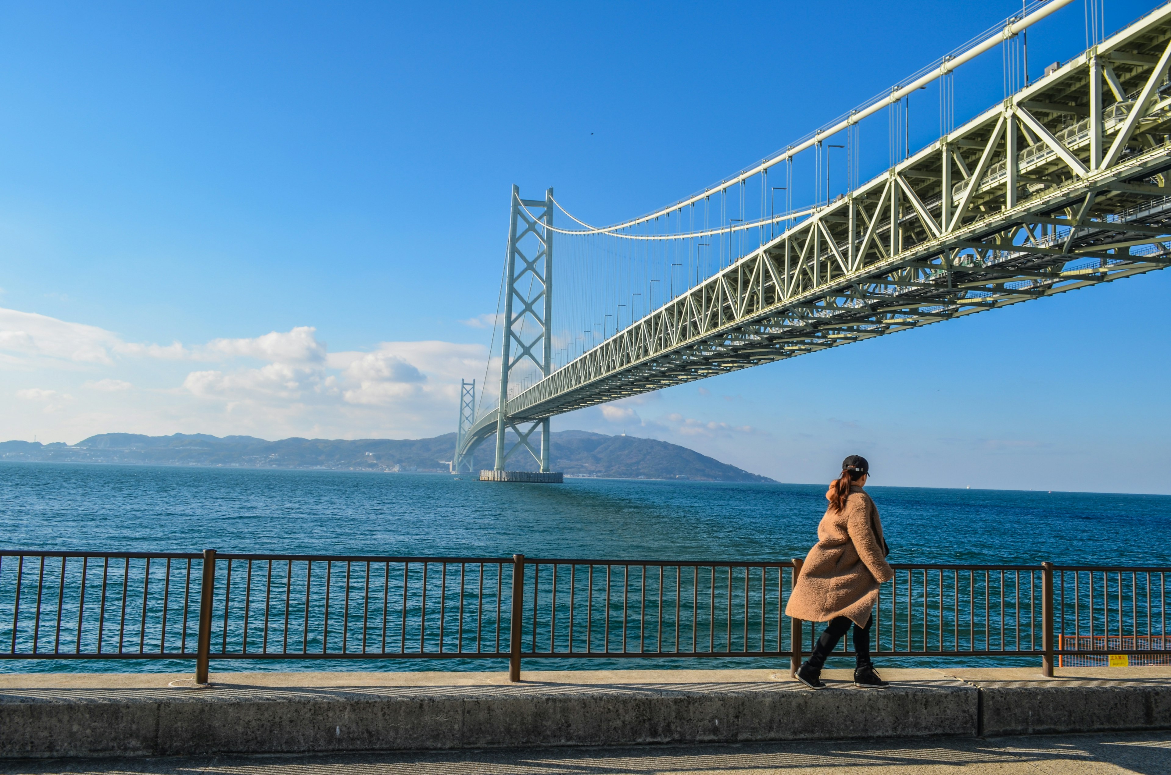 一名女性在海岸上行走，背景是明石海峡大桥