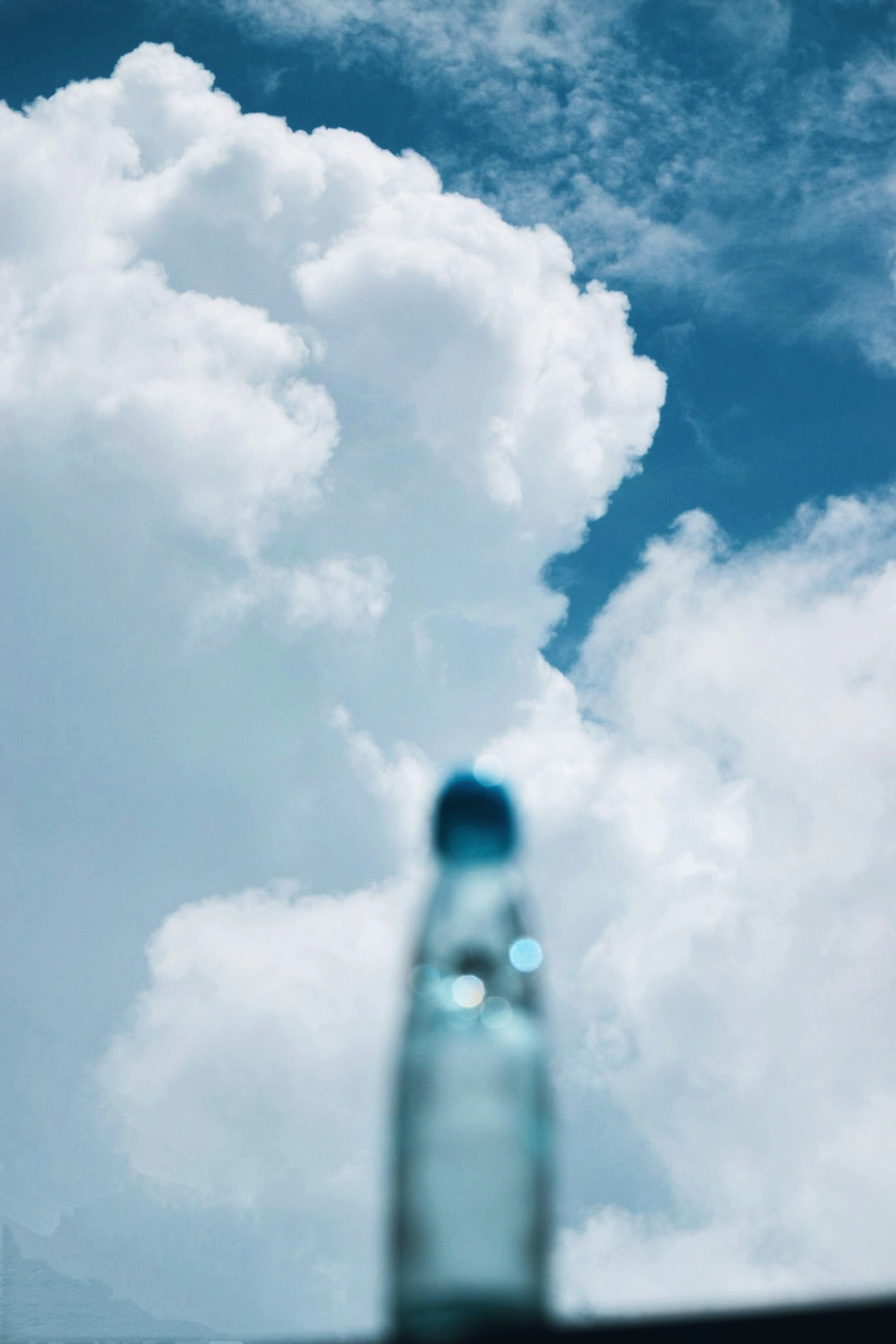 Verschwommene Abbildung einer Wasserflasche vor blauem Himmel und weißen Wolken