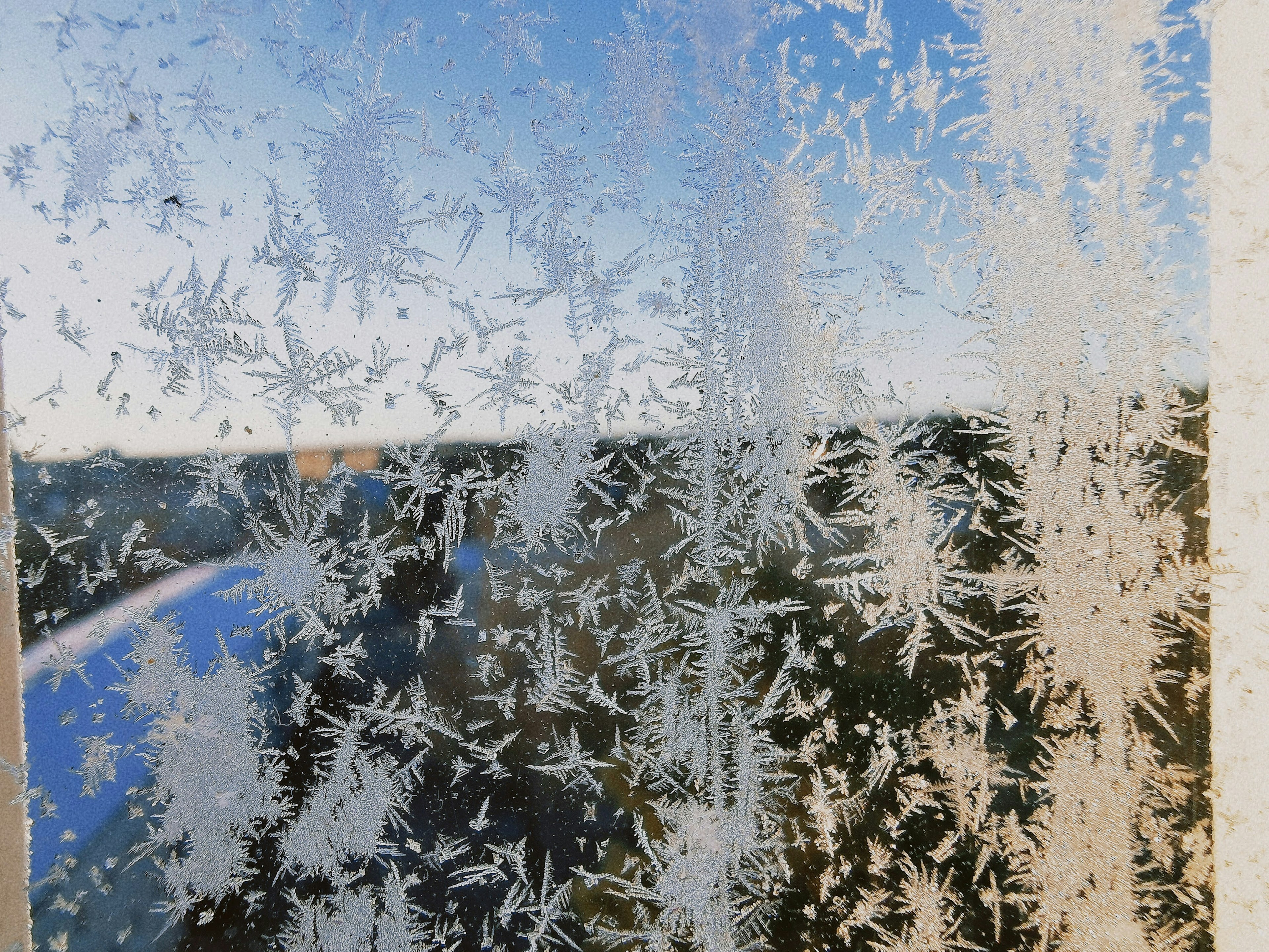 Eisige Fensterscheibe mit komplizierten Eiskristallen, die einen blauen Himmel und eine Landschaft zeigen