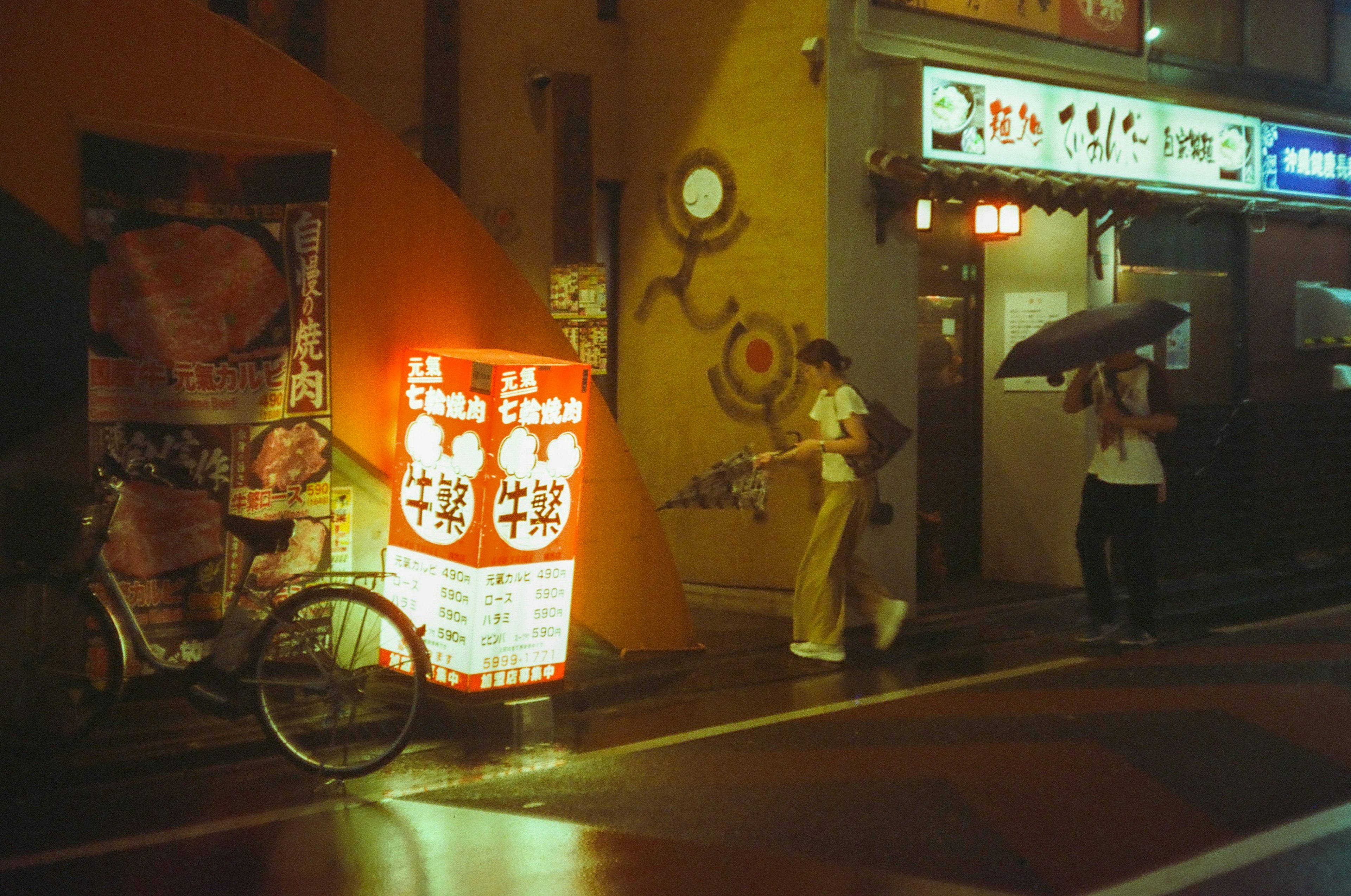 夜の街で傘をさした人と自転車のある風景 赤い看板が特徴的