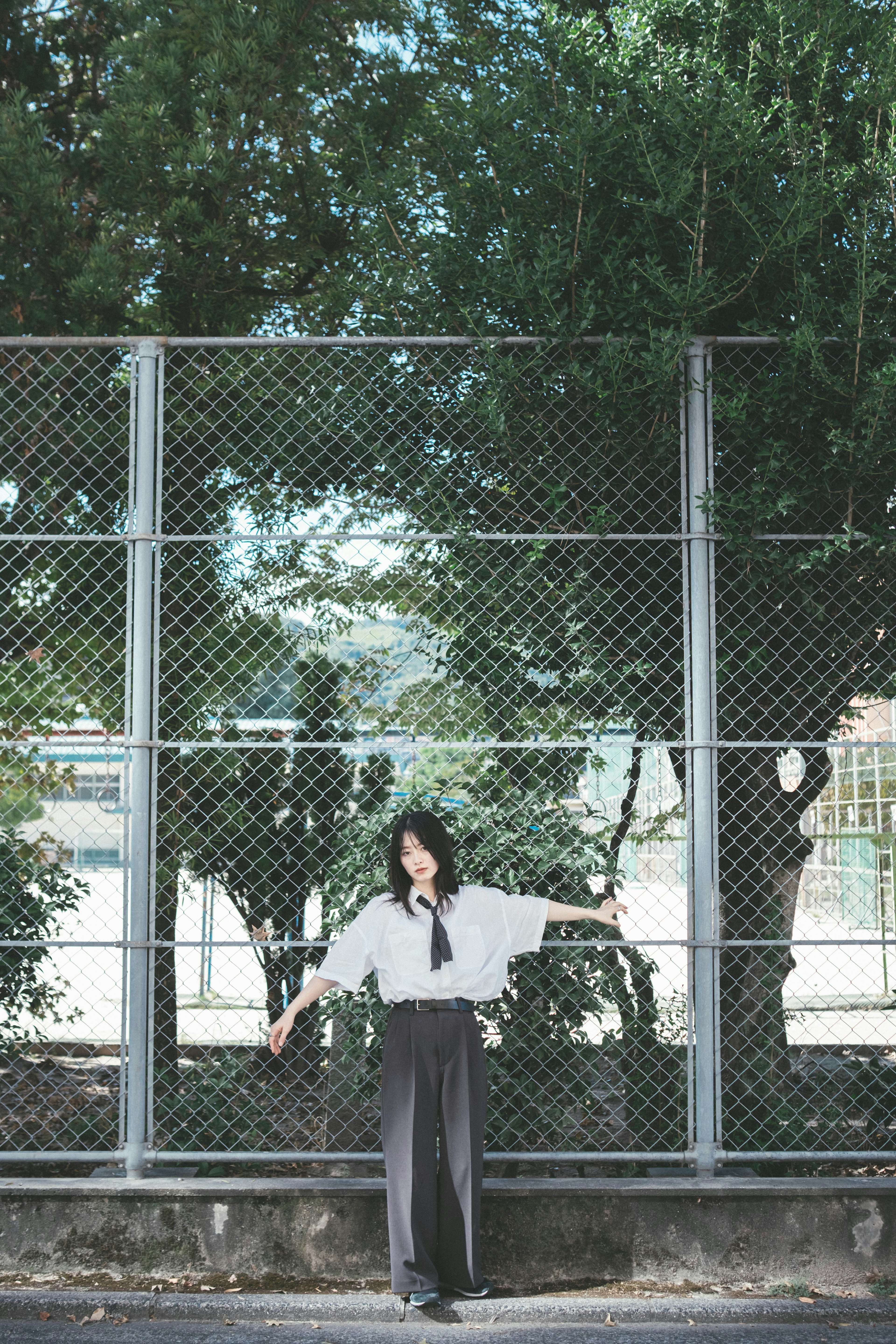 A woman stands in front of a fence wearing a white shirt and black pants surrounded by green trees