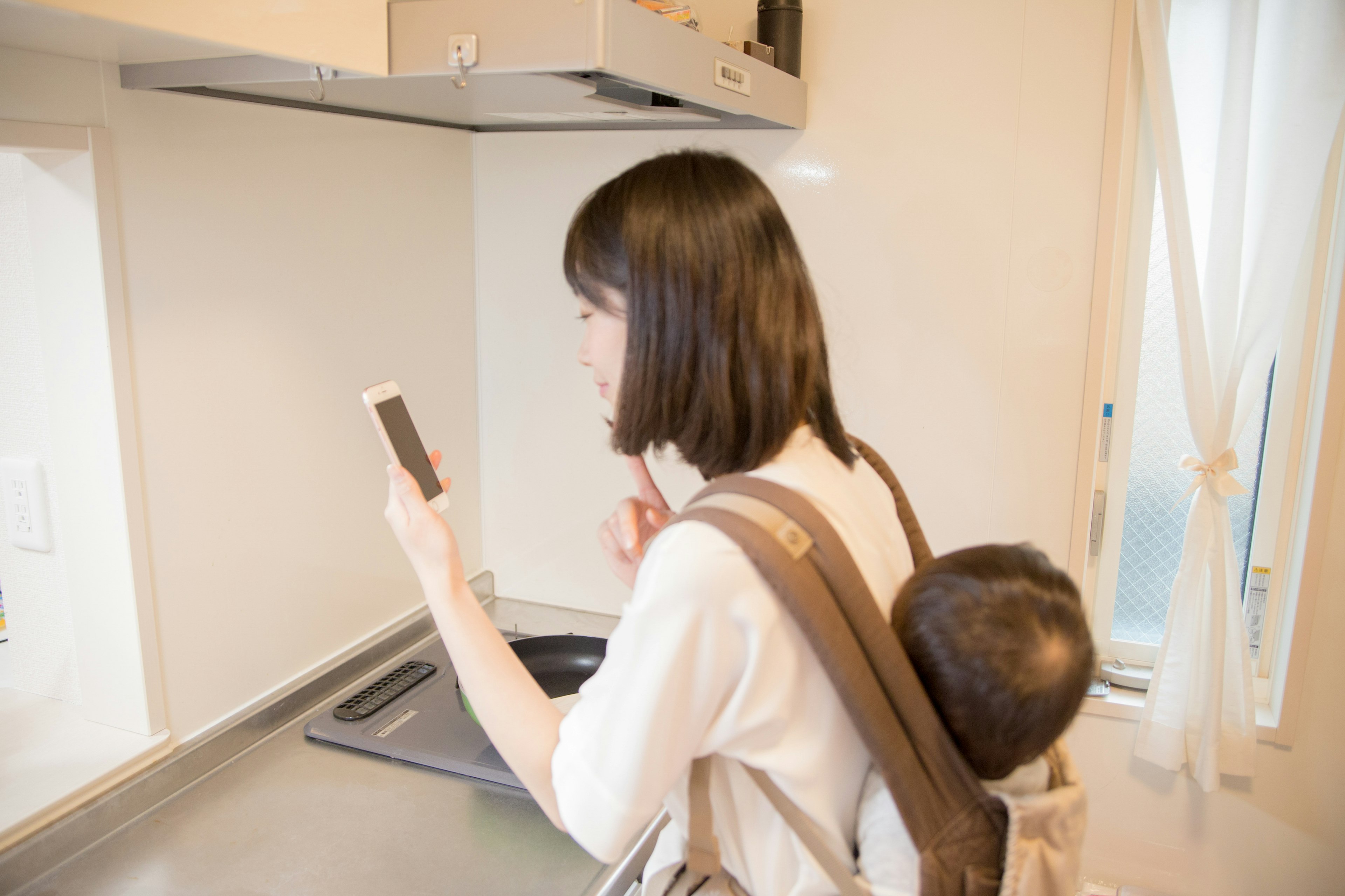 Madre usando un smartphone en la cocina con un niño en su espalda