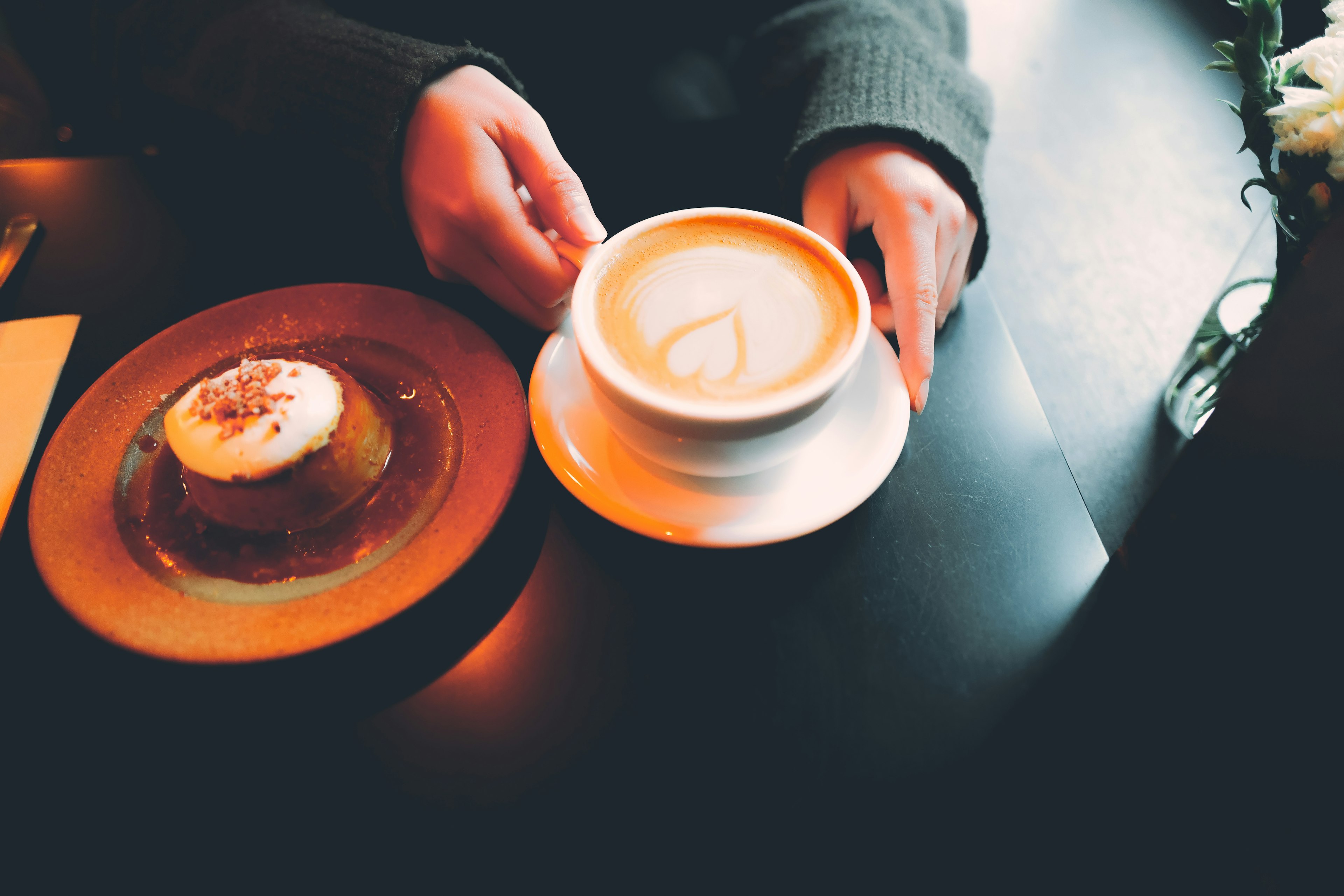 Mano sosteniendo un café con leche y un plato de postre en una mesa