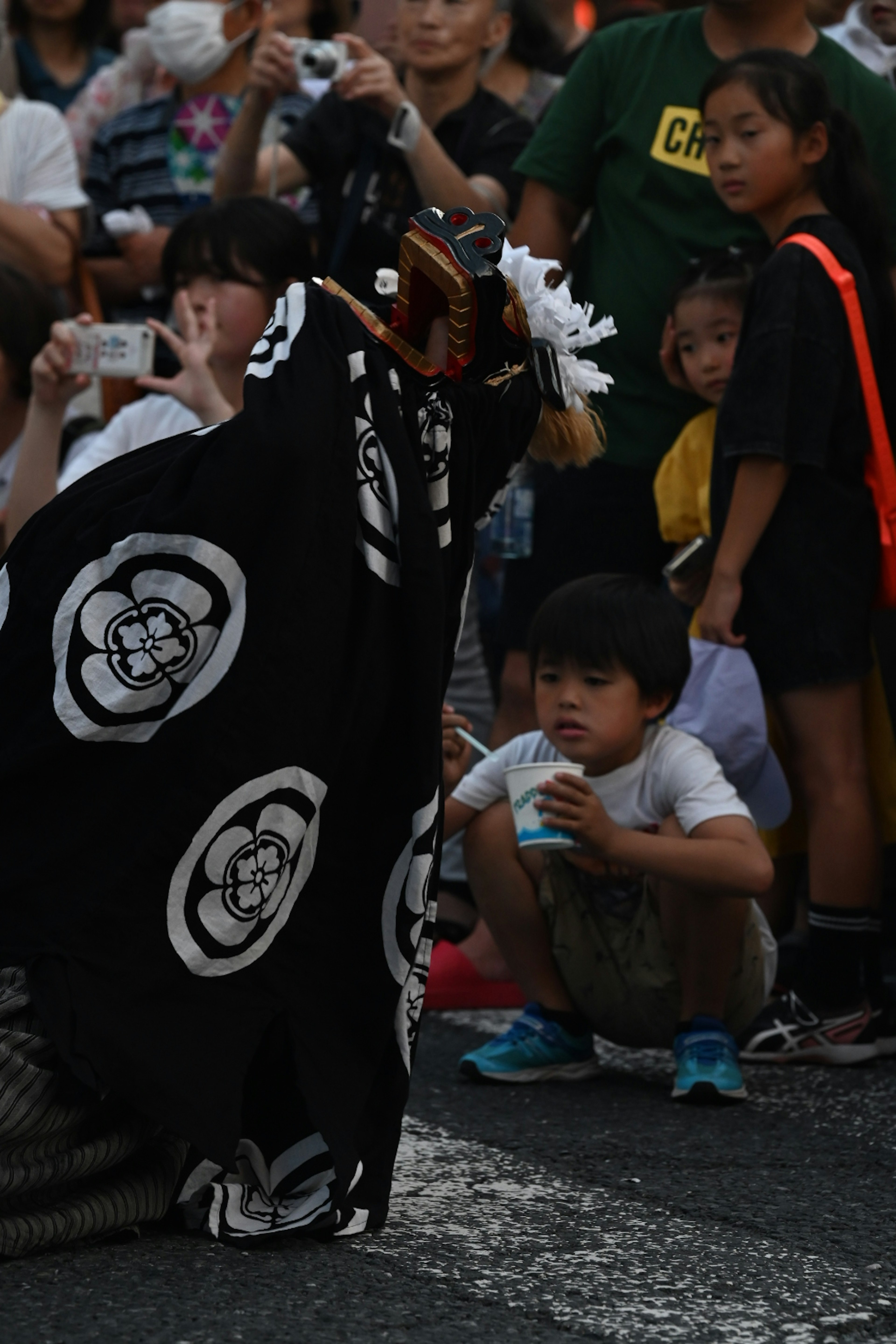 A traditional performer in a black outfit with white patterns entertaining a child sitting on the ground during a festival