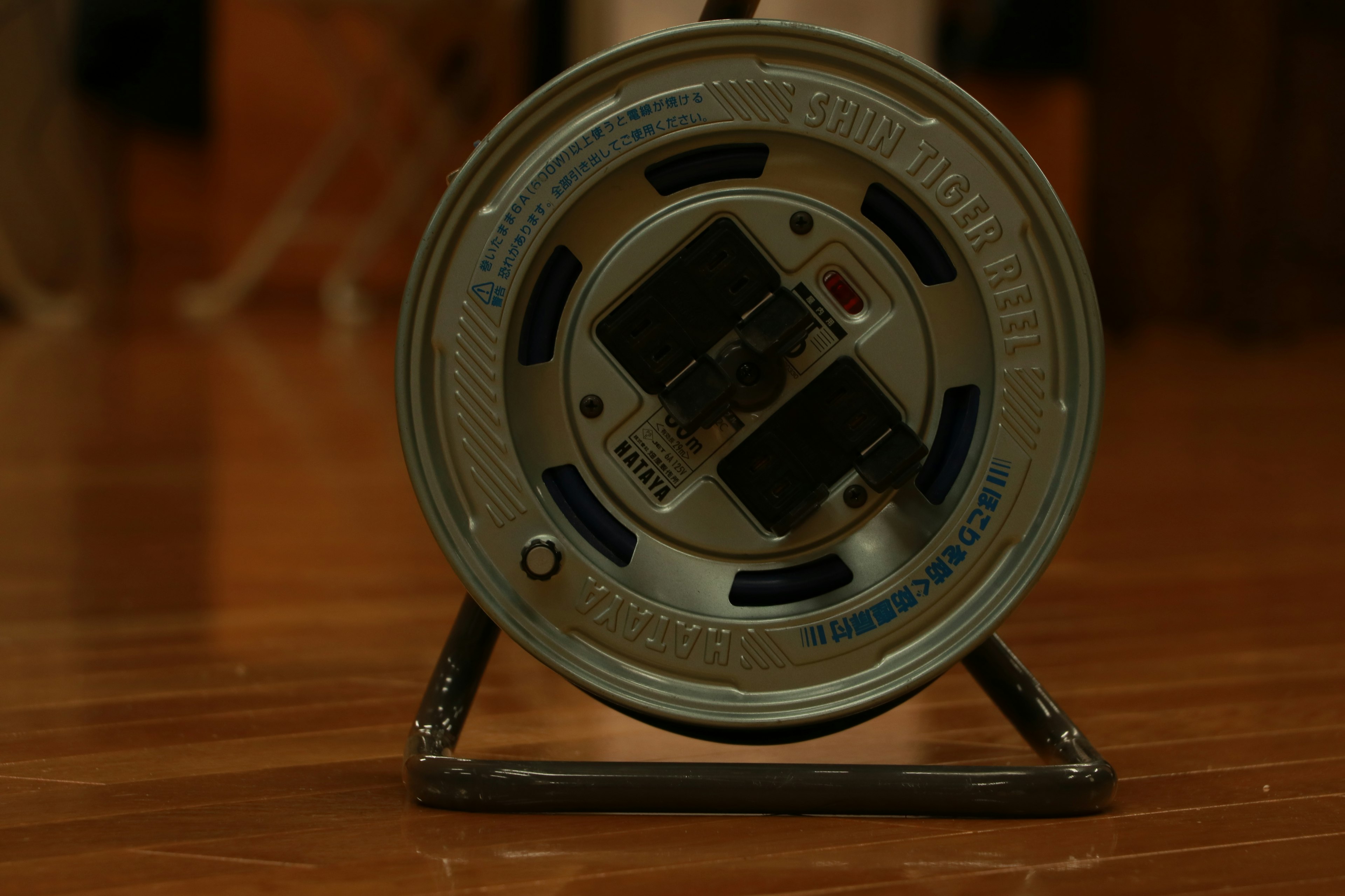 Image of a metallic reel placed on the floor featuring black switches on top and a wooden floor background