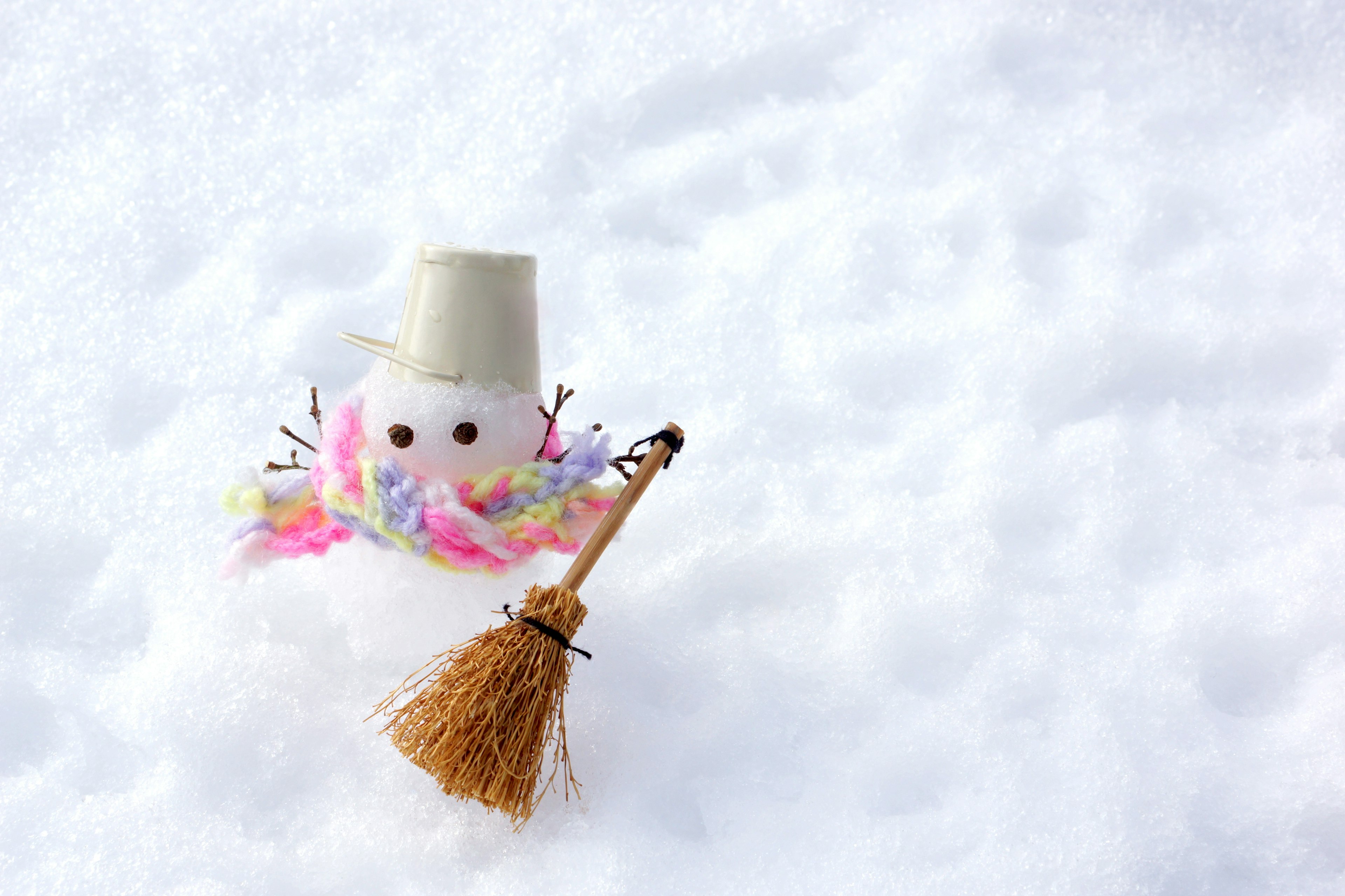 A small snowman in the snow wearing a colorful scarf and a light-colored hat holding a broom