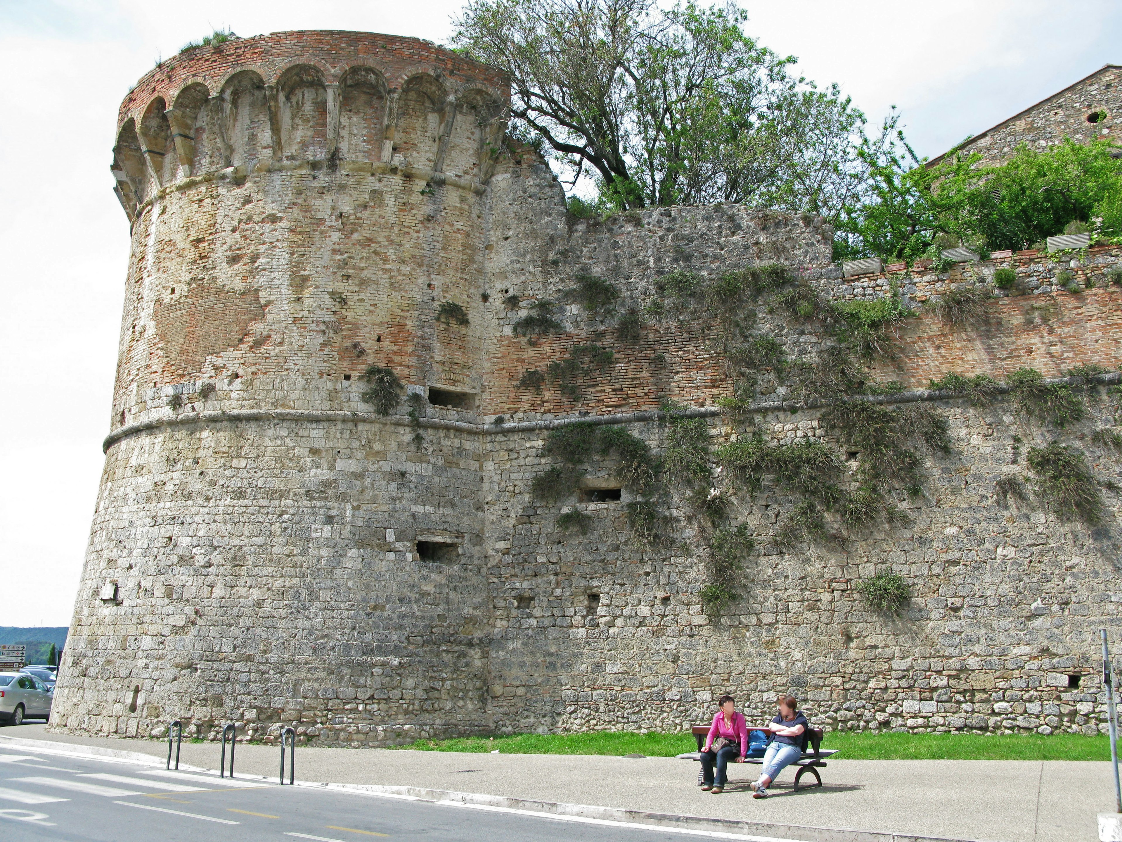 Muro di pietra storico con una torre rotonda e due persone sedute su una panchina