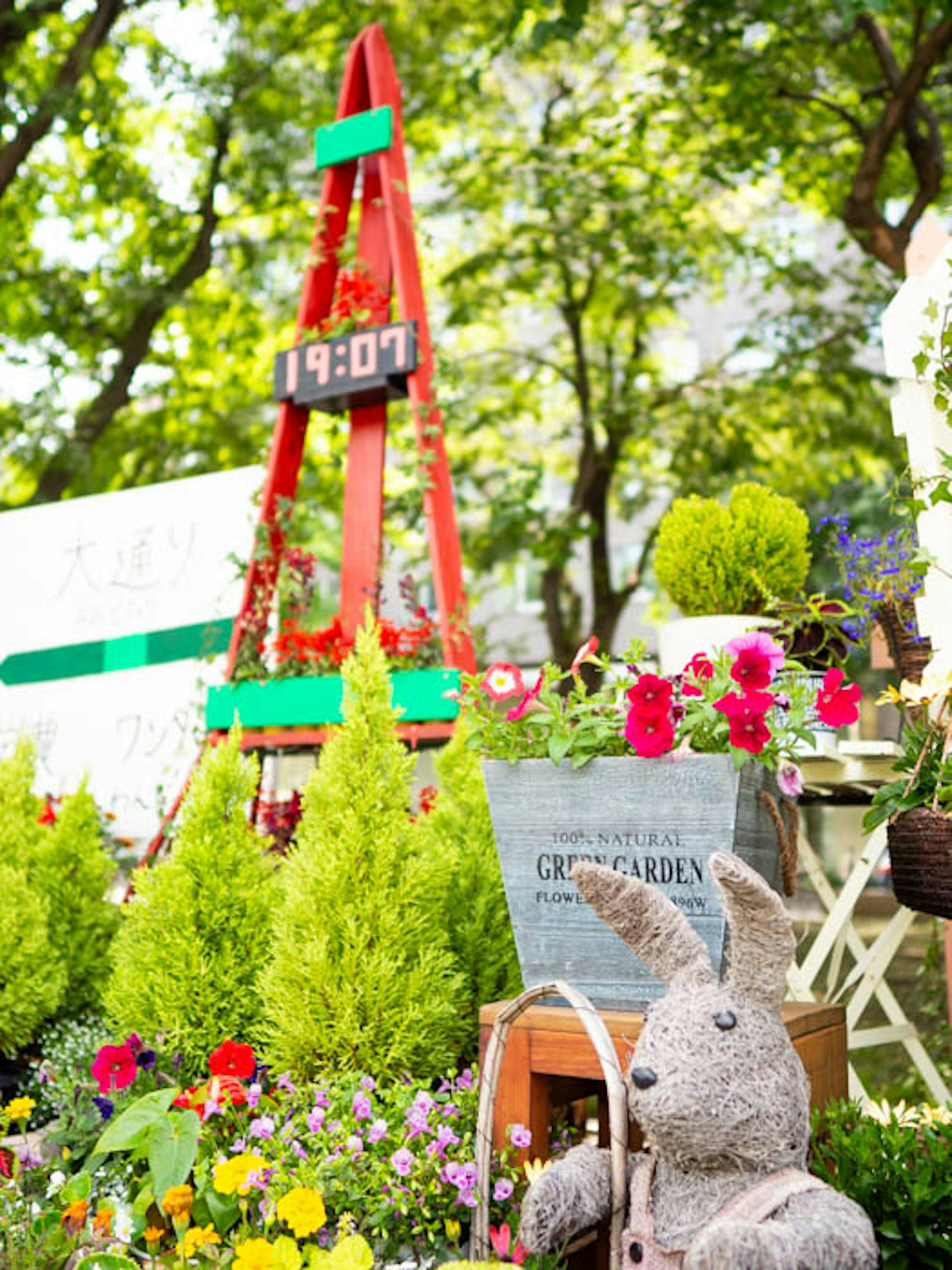 Flores coloridas y arbustos verdes rodean una escena de jardín con una estructura de reloj rojo y un juguete de peluche de conejo