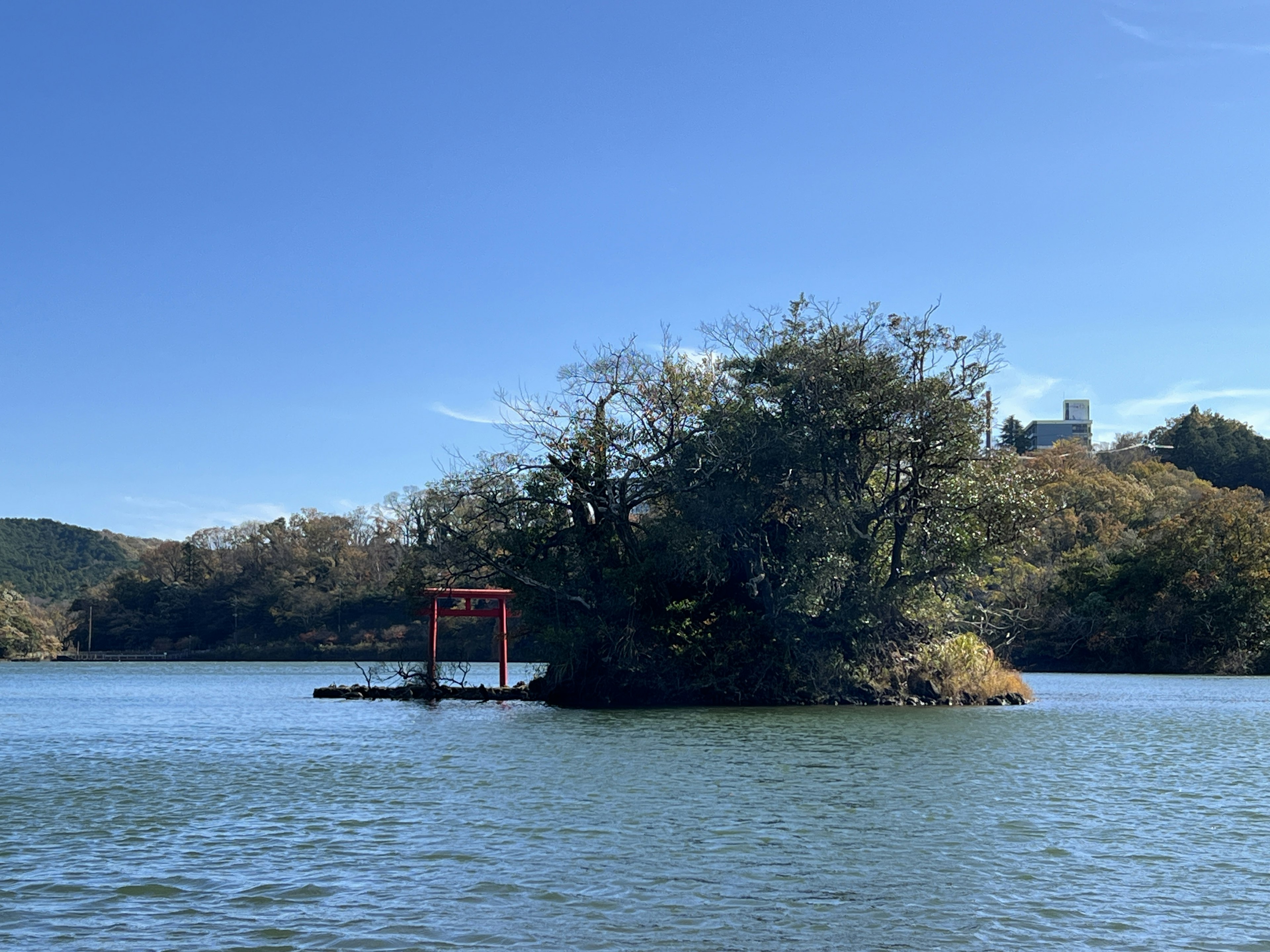 Pulau kecil dengan gerbang torii merah dikelilingi oleh air tenang dan langit biru