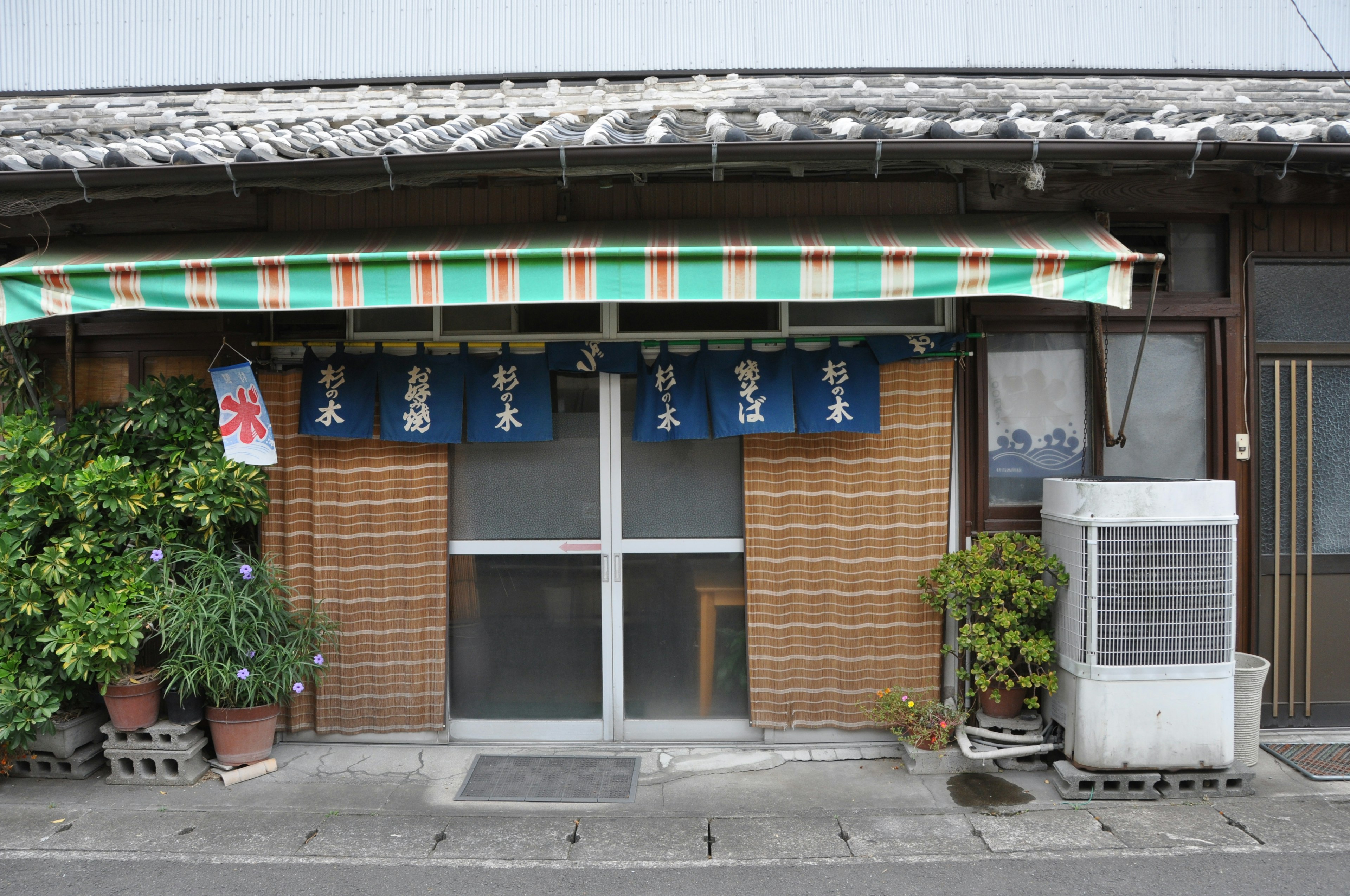 Traditional Japanese shop exterior with striped awning and blue noren visible plants and air conditioning unit