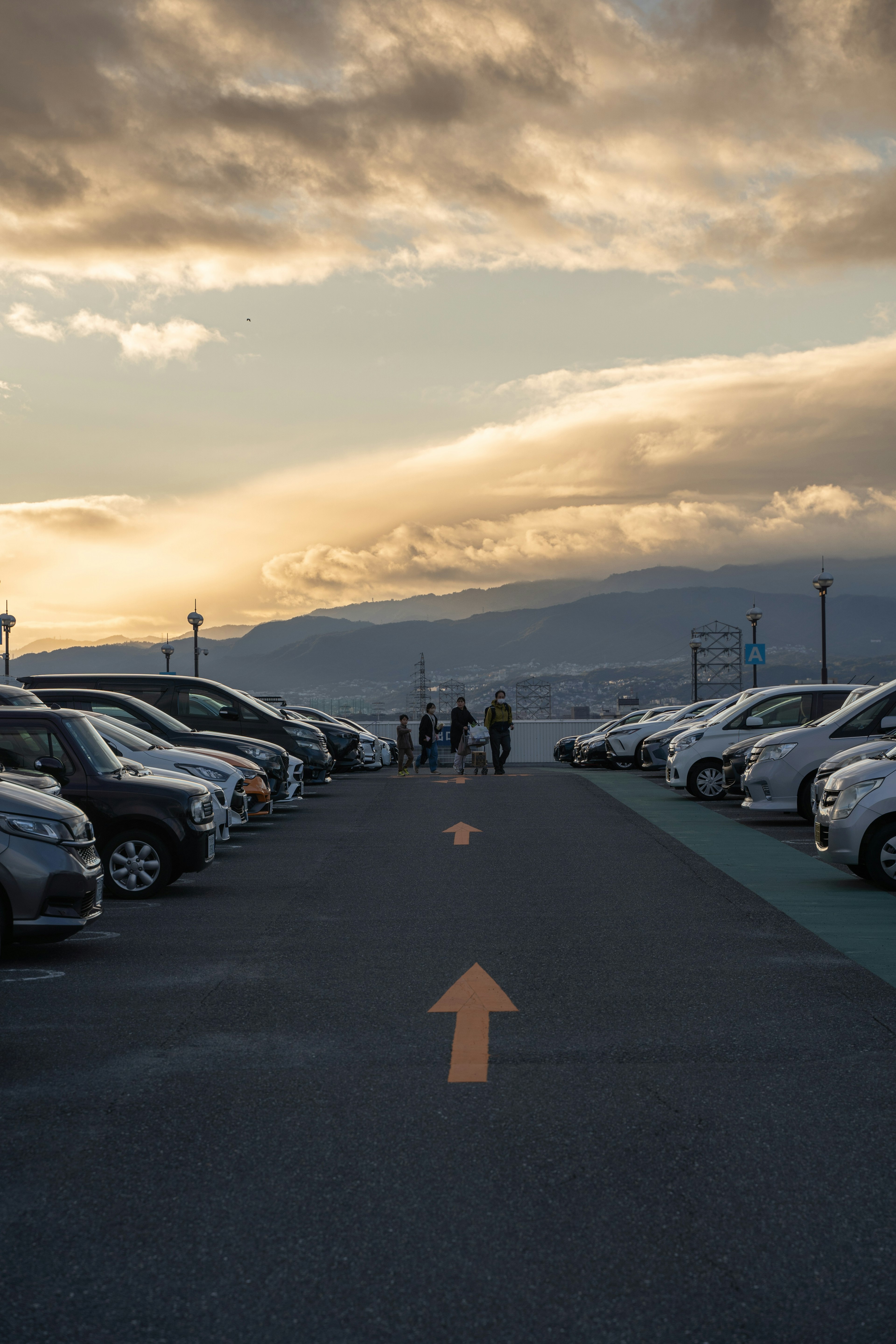 駐車場の車が並び、夕焼けの空の下に人々が歩いている