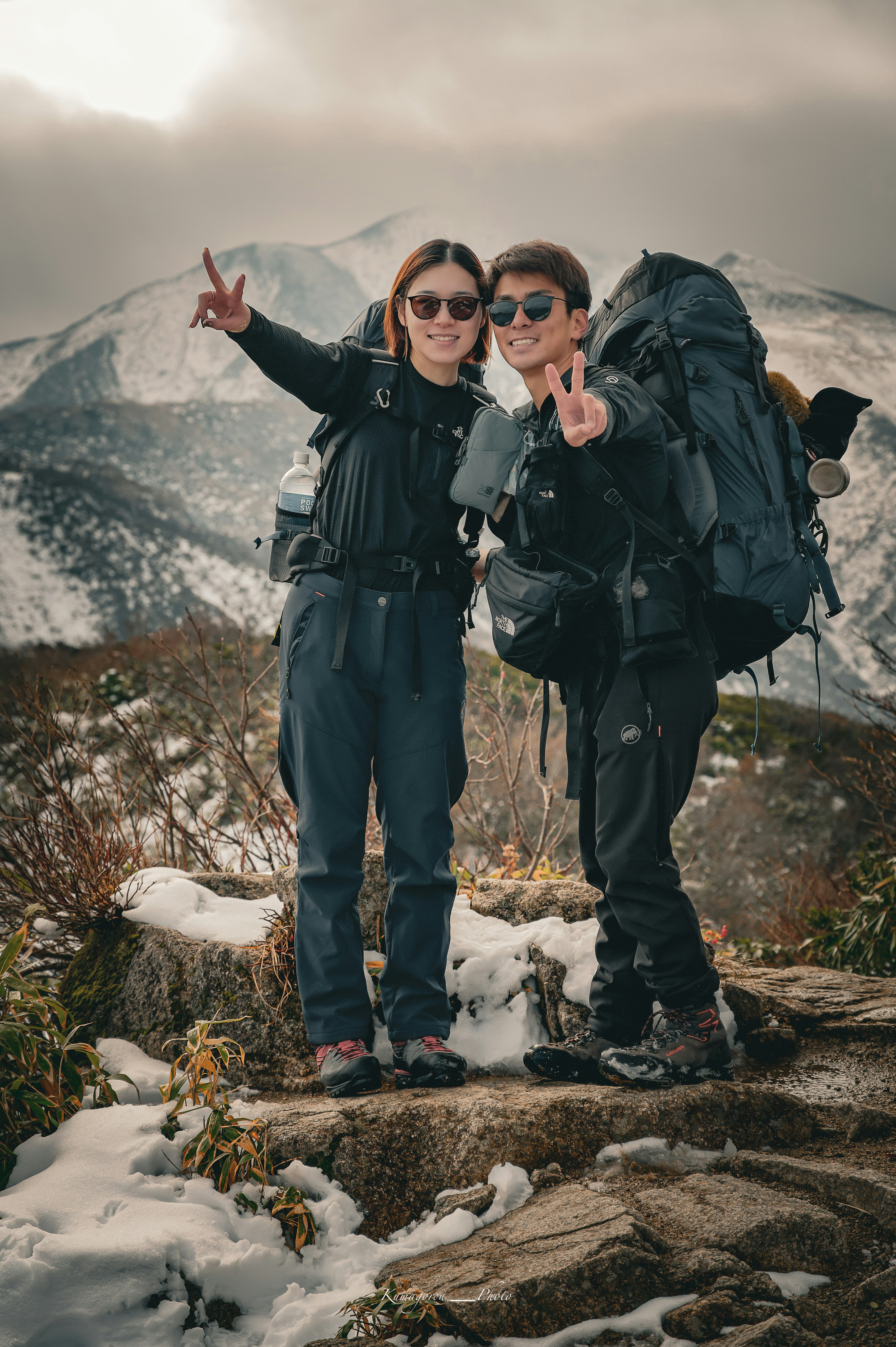 二人の登山者が山の頂上でポーズをとっている背景には雪をかぶった山々が広がる