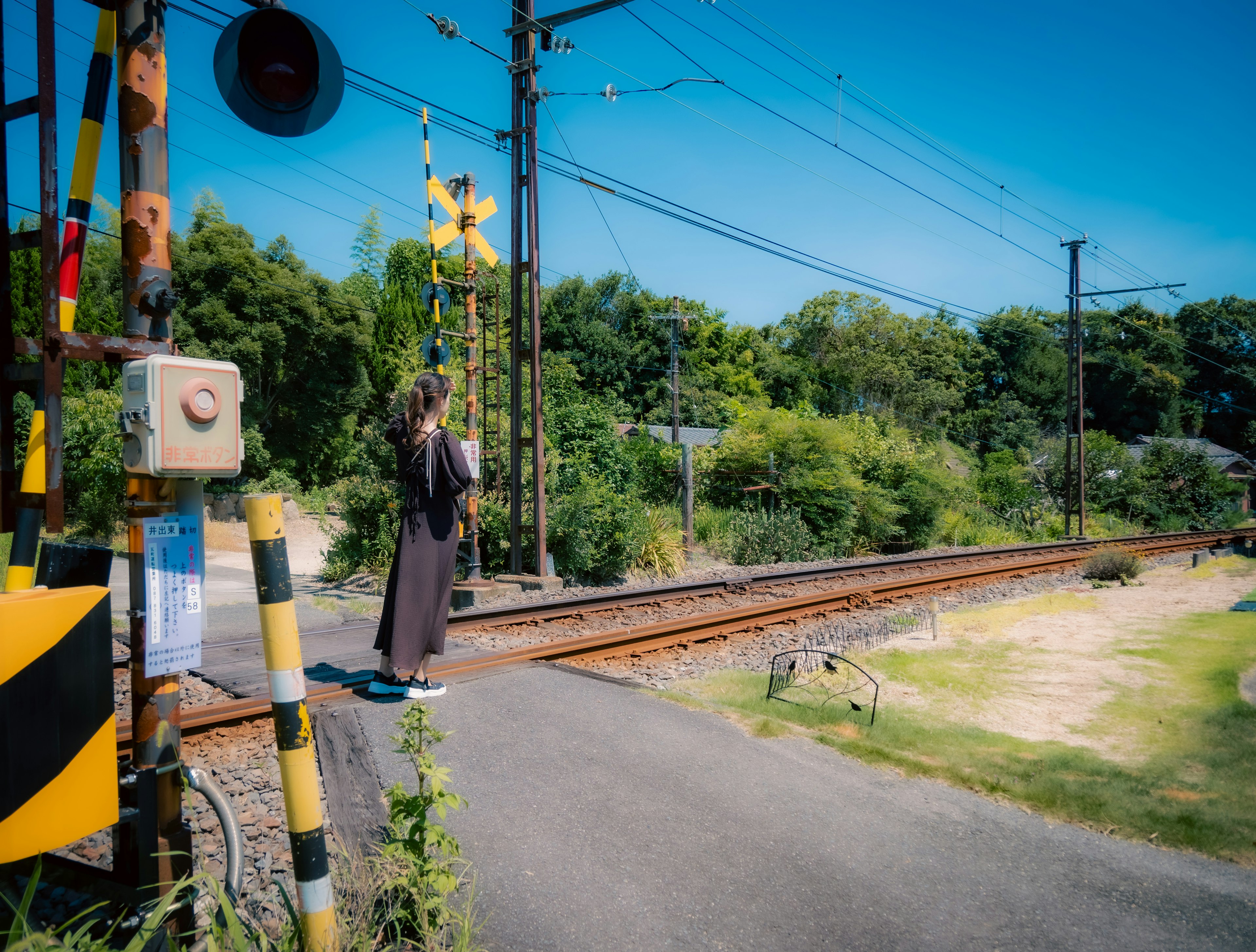 Person standing at a railway crossing with lush greenery in the background on a sunny day