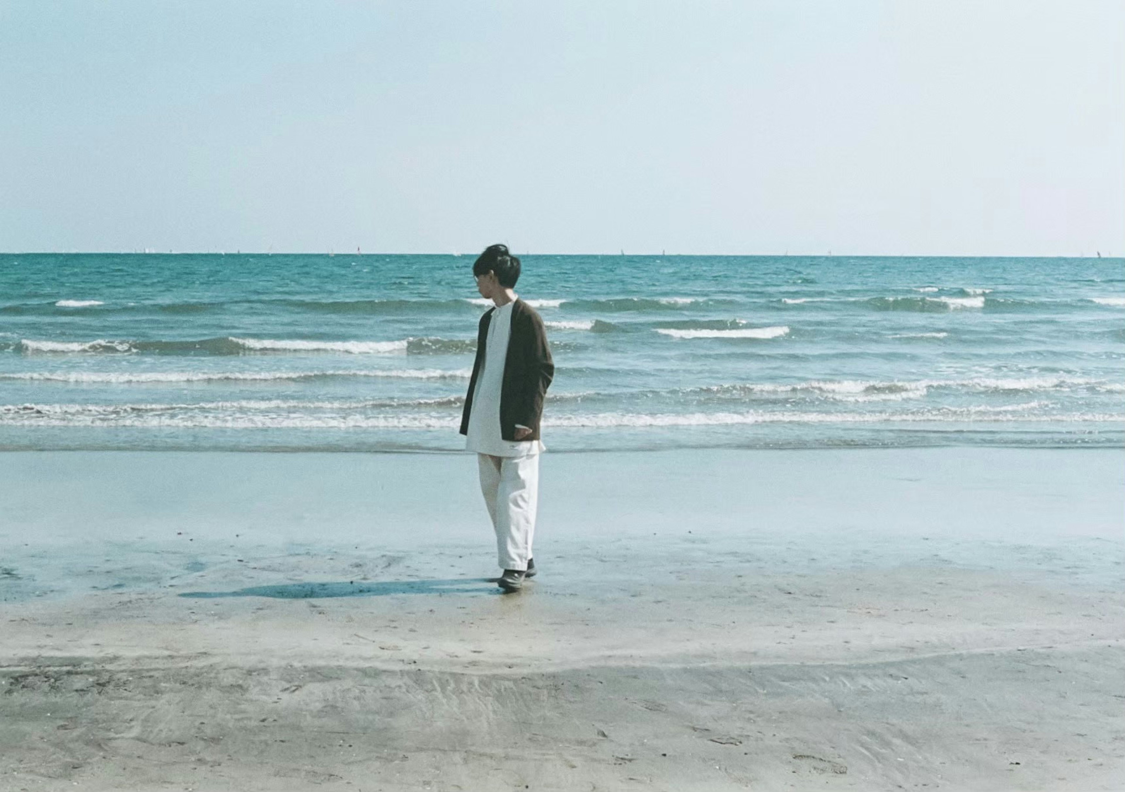 Ein Mann steht am Meer in weißen Hosen und einer grauen Jacke mit blauem Meer und Wellen