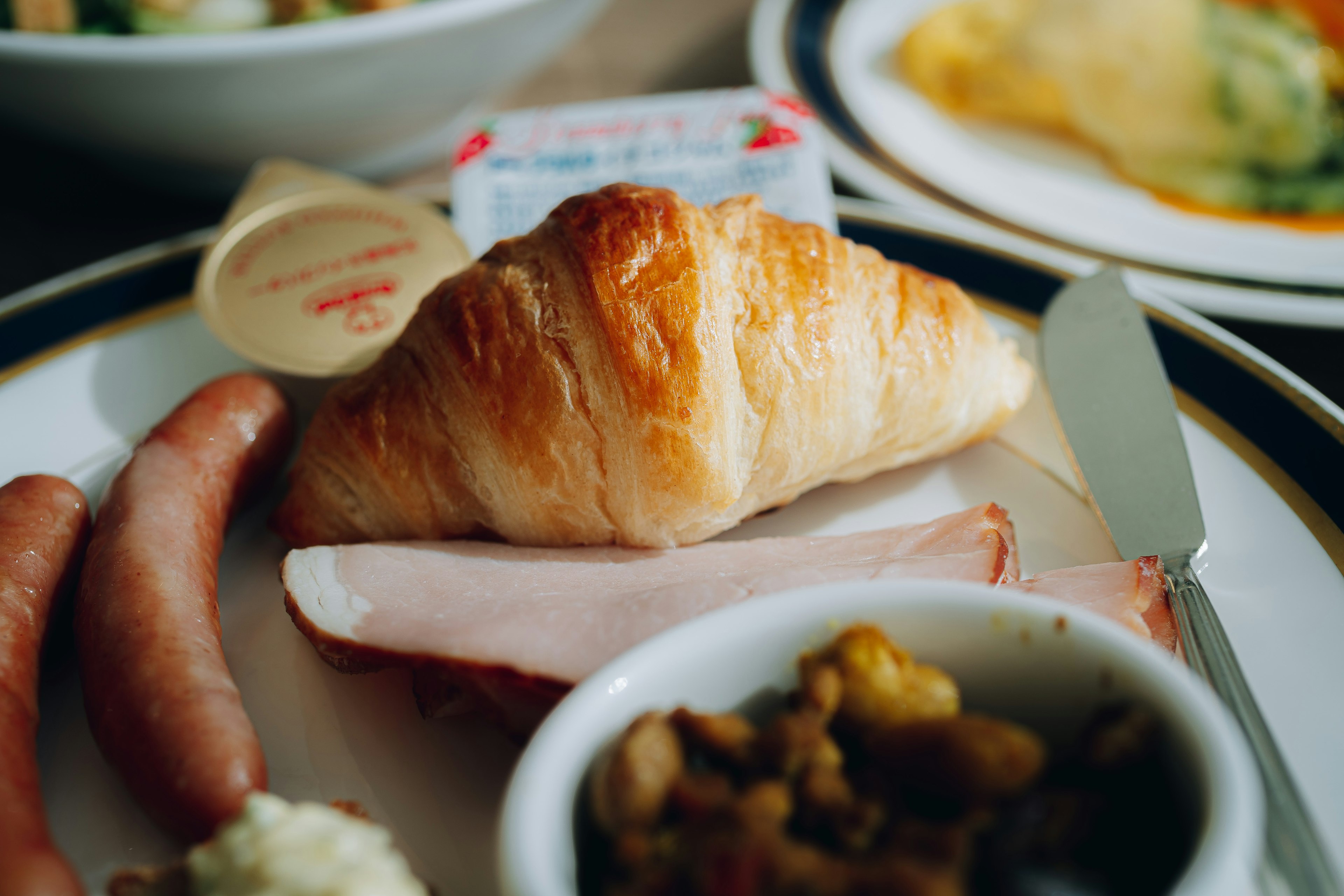 Frühstücksteller mit Croissant Würstchen Schinken und einer kleinen Schüssel mit gemischtem Gemüse