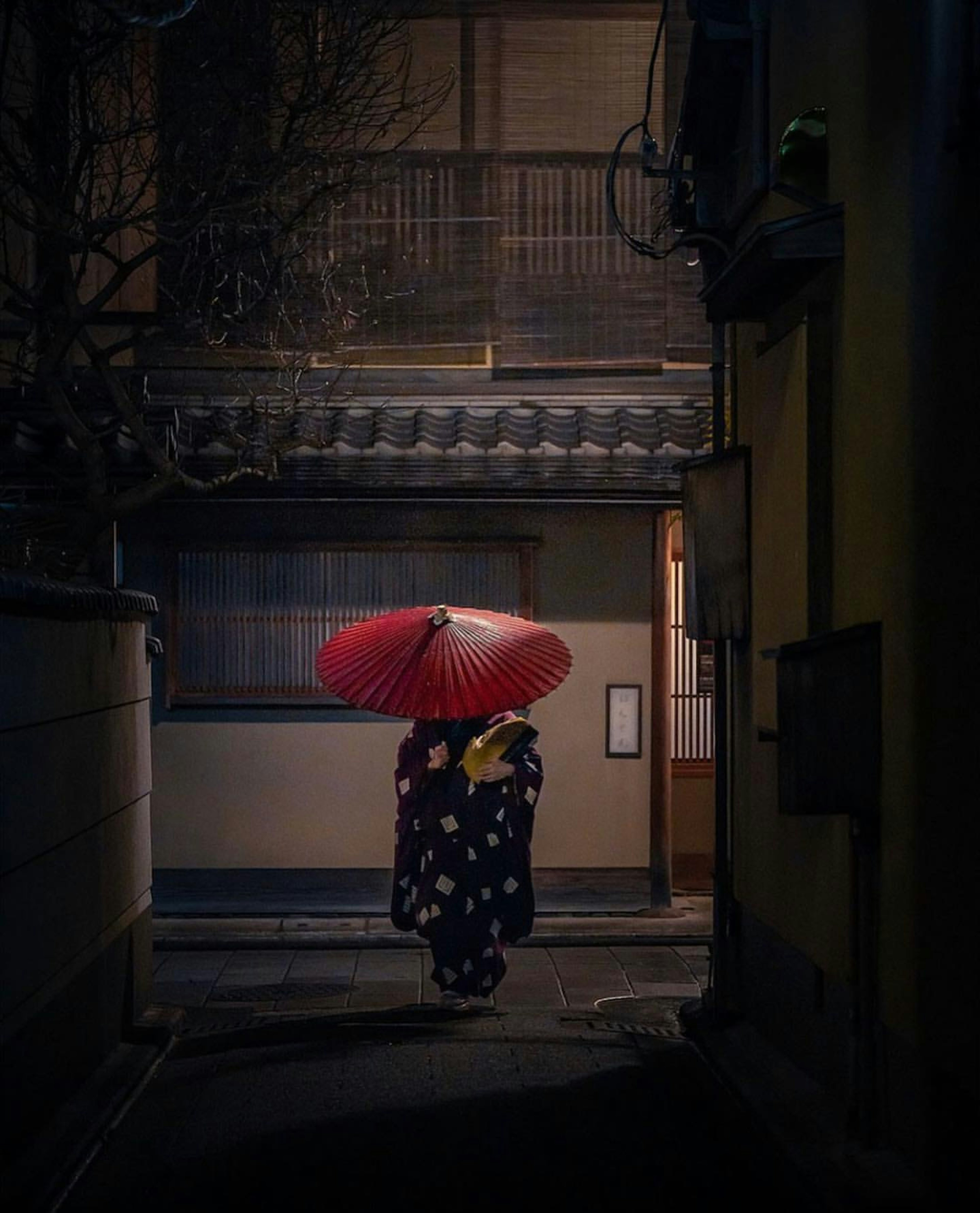 Une femme en kimono portant un parapluie rouge marchant dans une ruelle nocturne