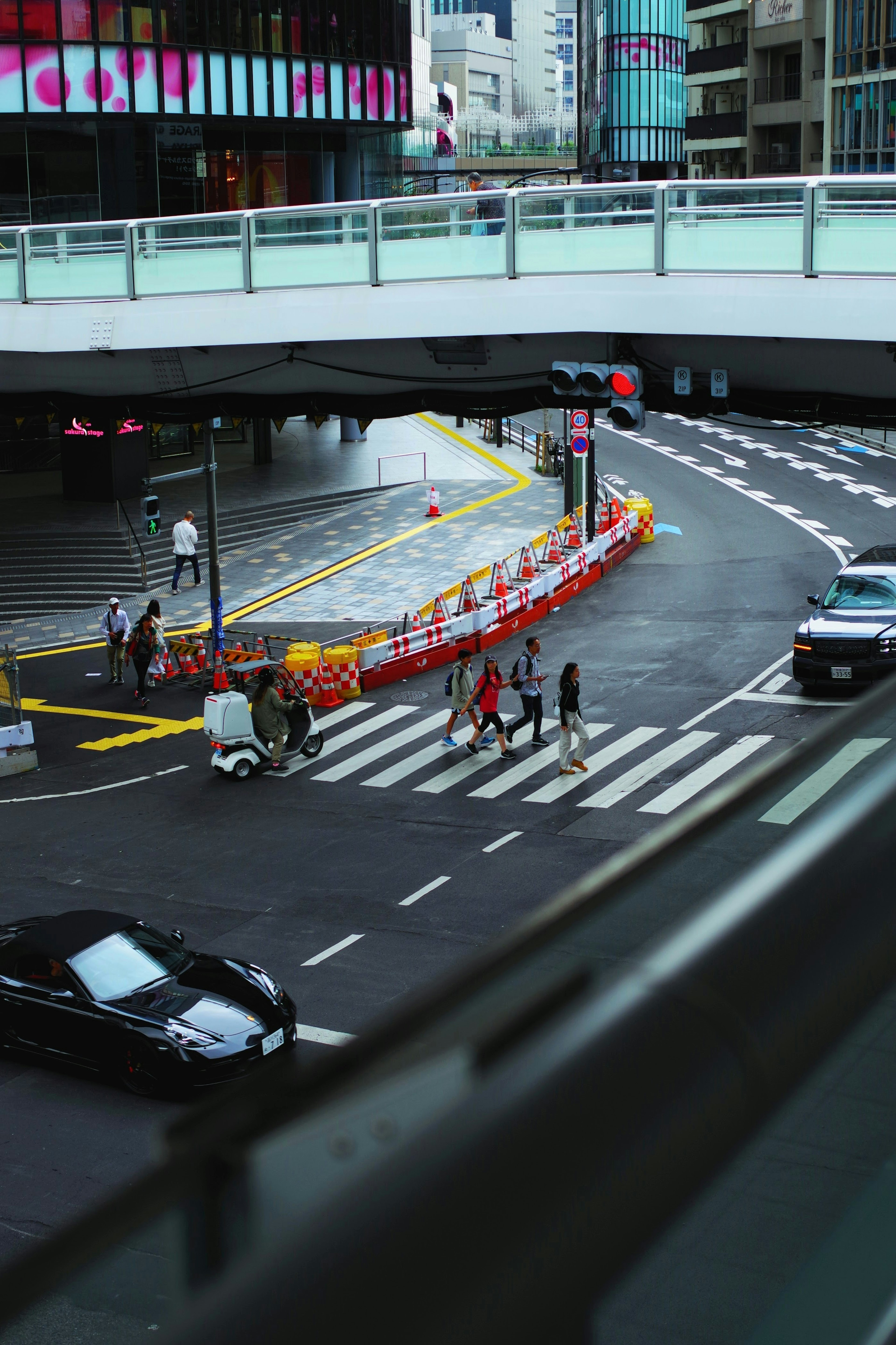 都市の交差点で歩行者と車両が行き交う様子 赤いバリケードと横断歩道が見える