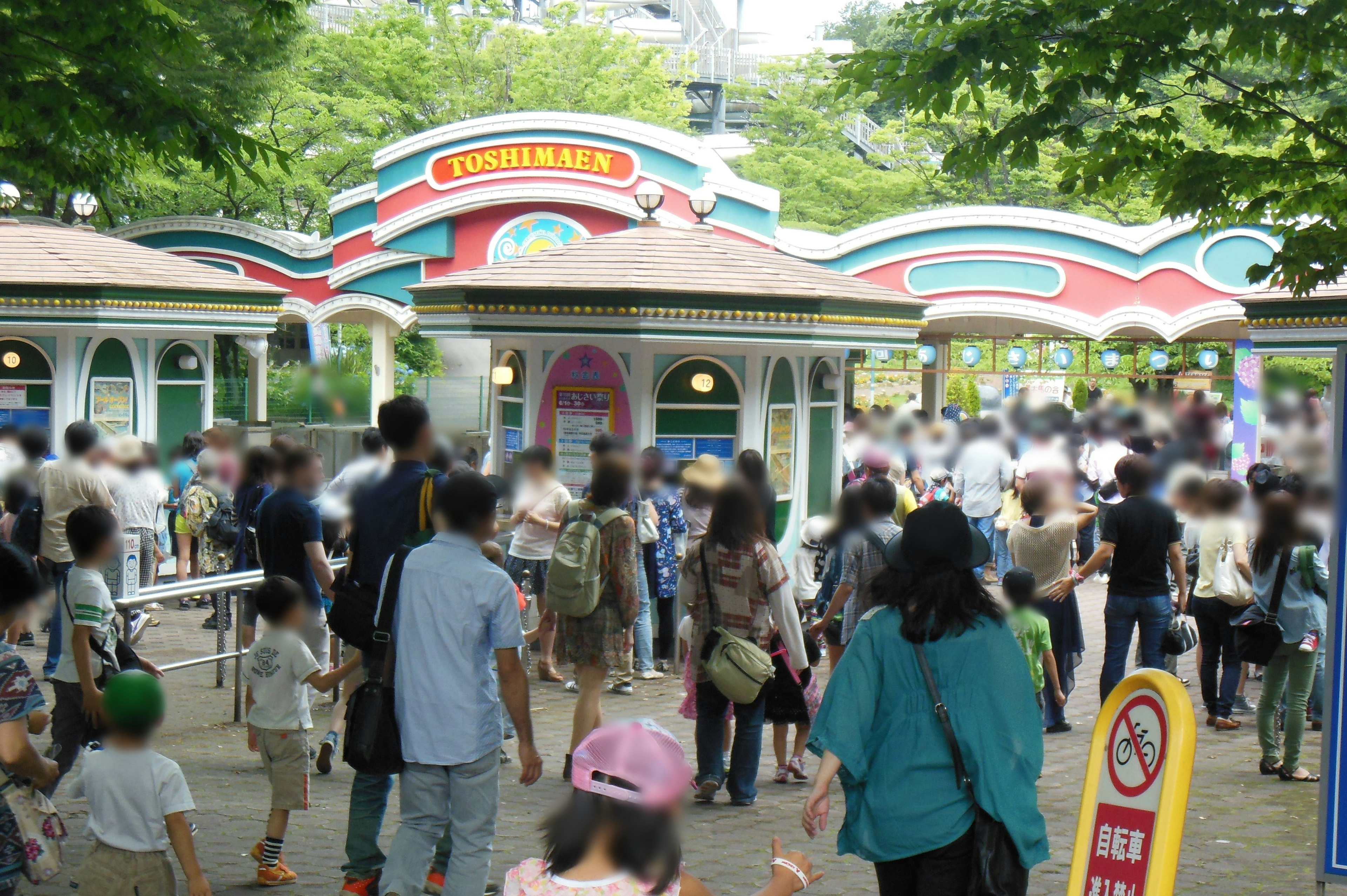 賑やかな遊園地の入り口で人々が行き交う様子 緑豊かな木々とカラフルな建物が特徴