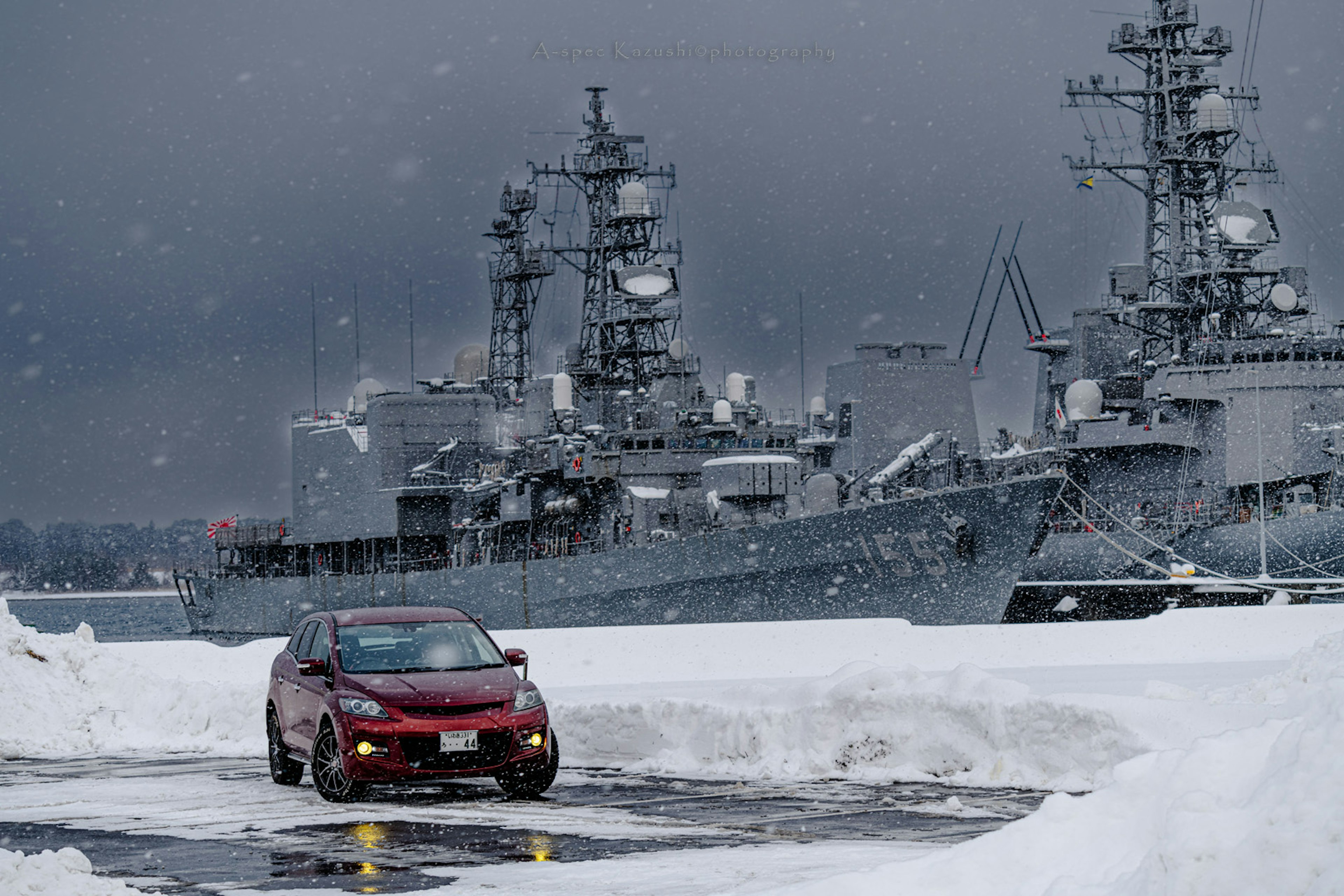 紅色汽車在雪中行駛，背景是海軍艦艇