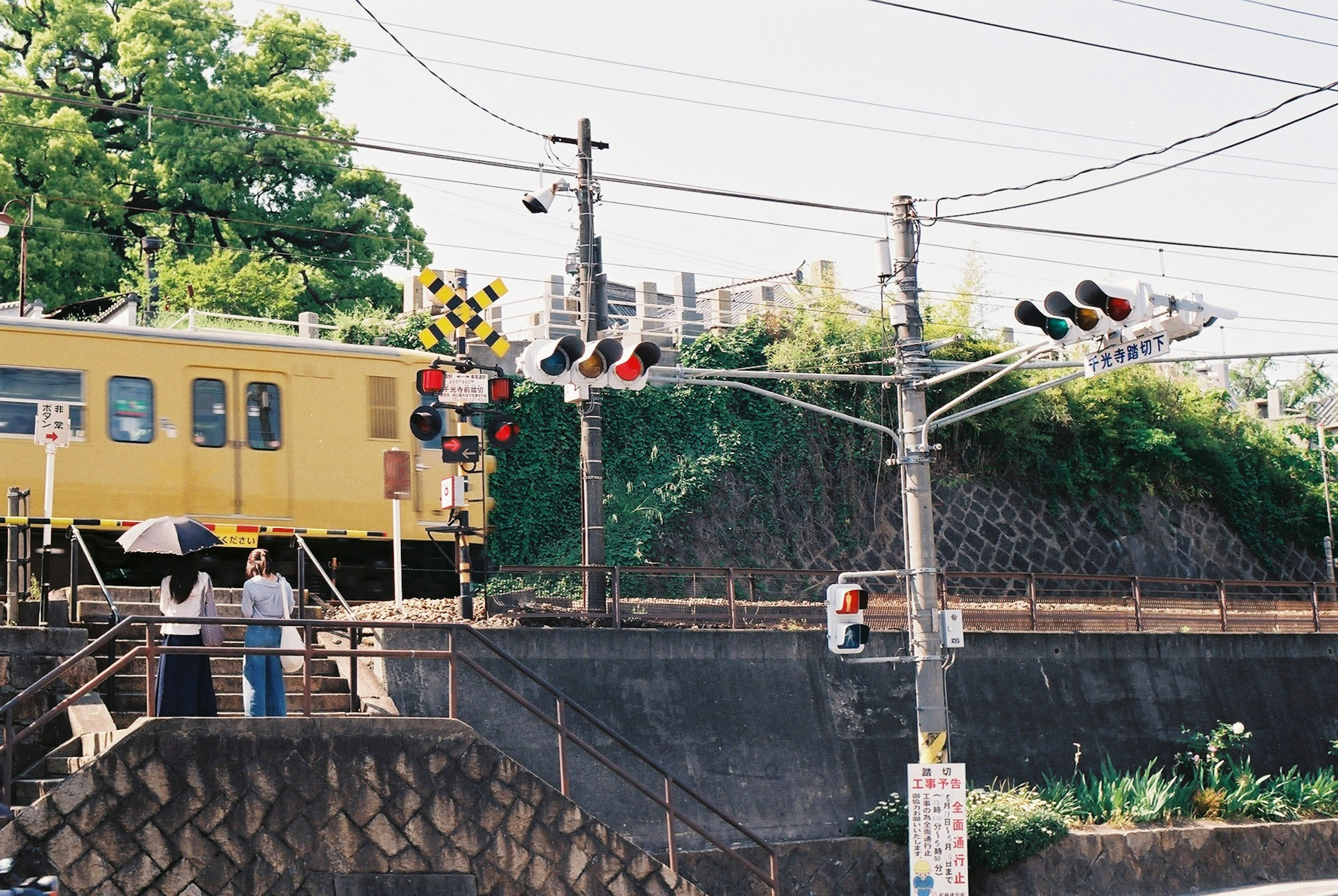 黄色い電車が通過する踏切の風景 人々が傘をさして待っている