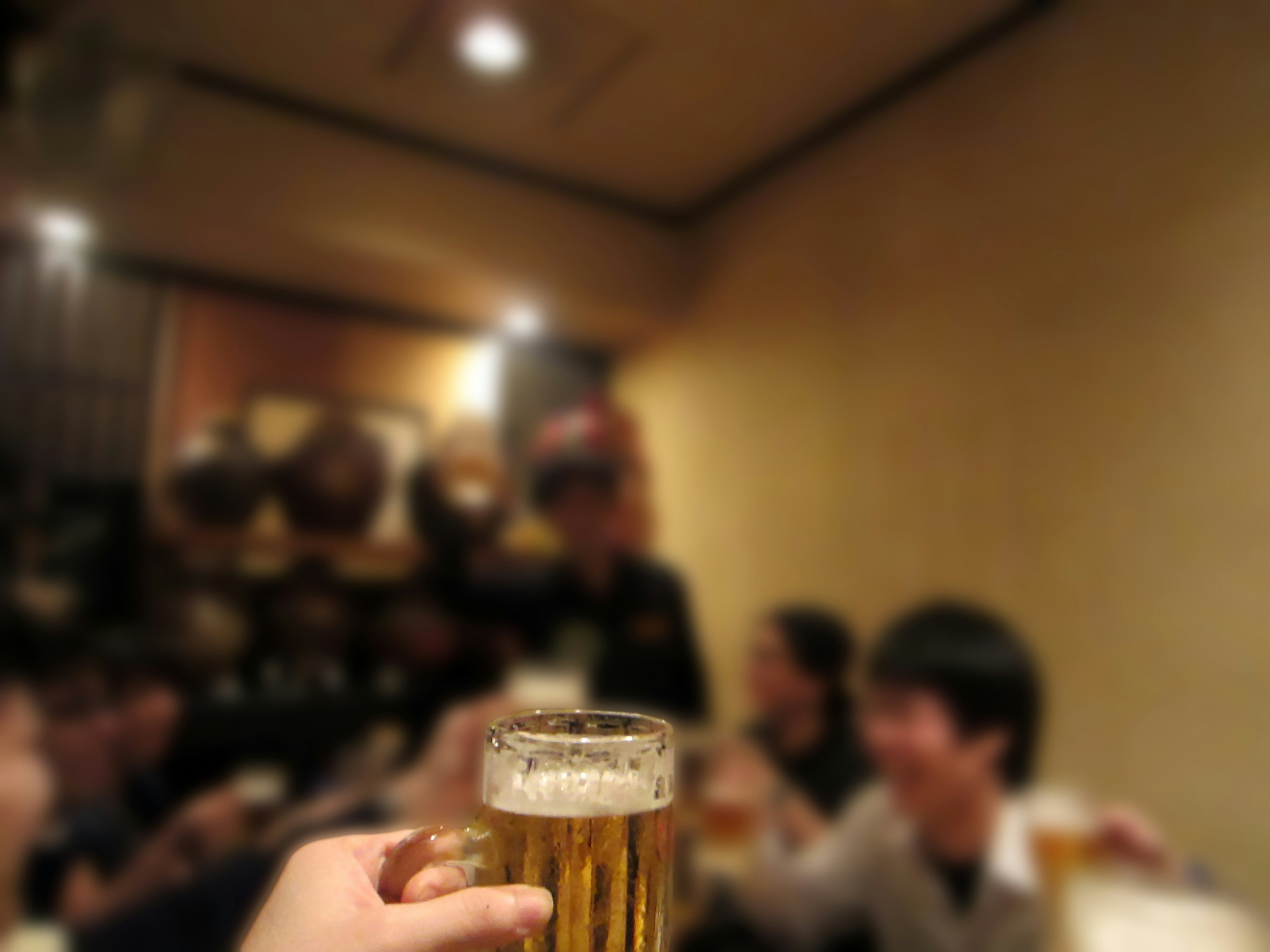 A hand holding a beer glass with people in the background at an izakaya