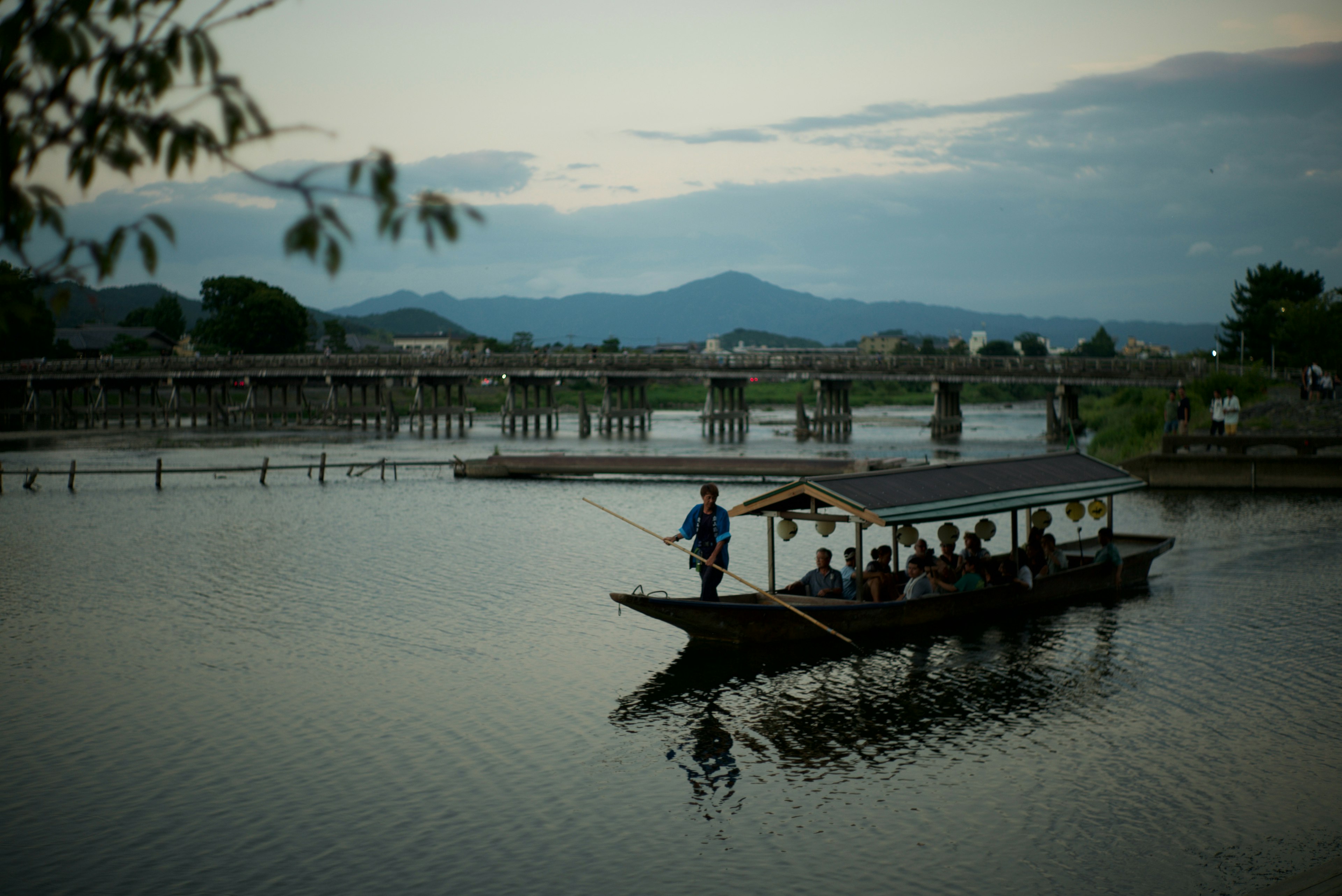 ทิวทัศน์ที่สวยงามของผู้คนในเรือในแม่น้ำที่สงบพร้อมสะพานและภูเขาในพื้นหลัง
