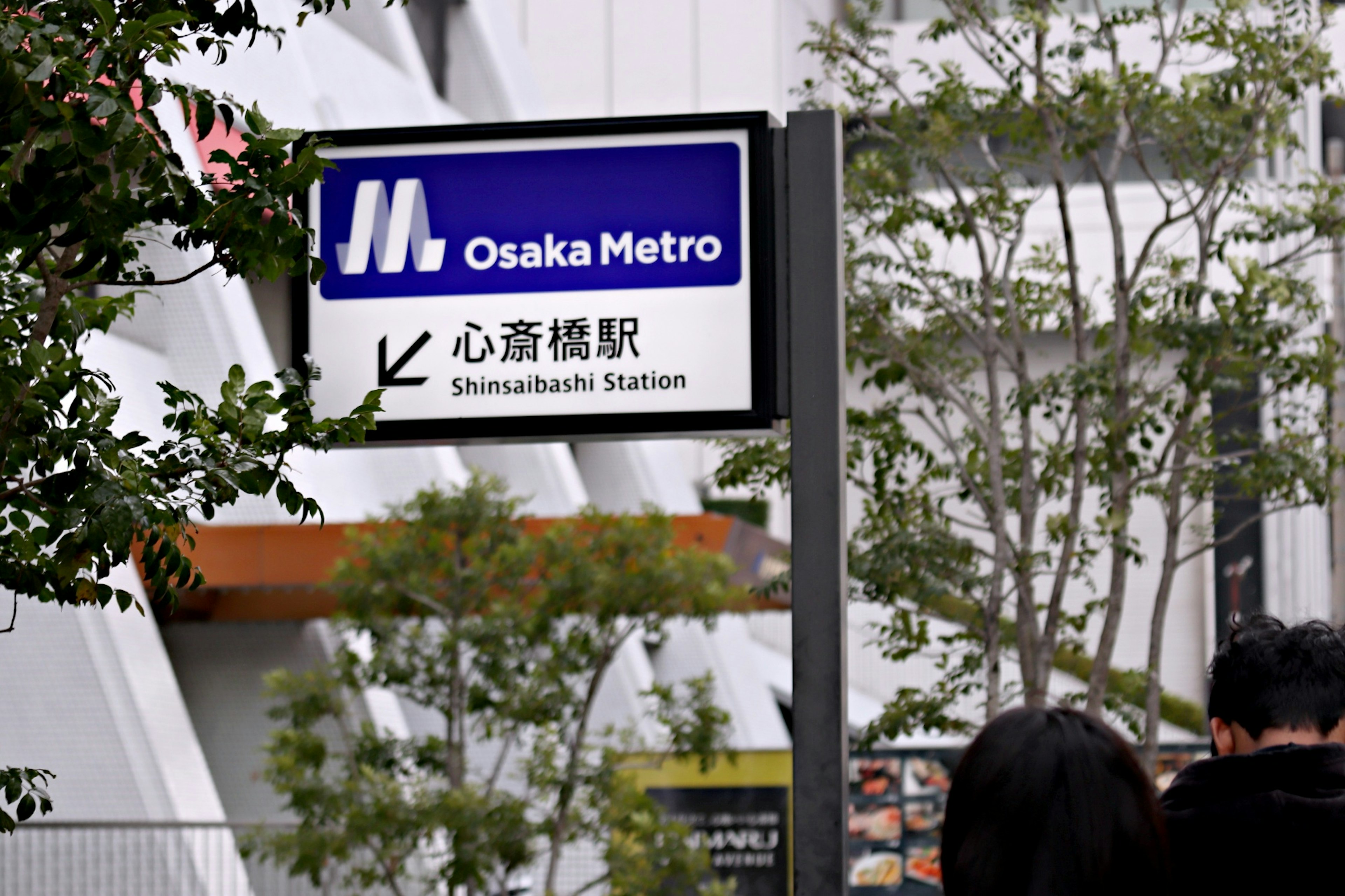 Papan stasiun Shinsaibashi dari Osaka Metro dengan vegetasi di sekitarnya