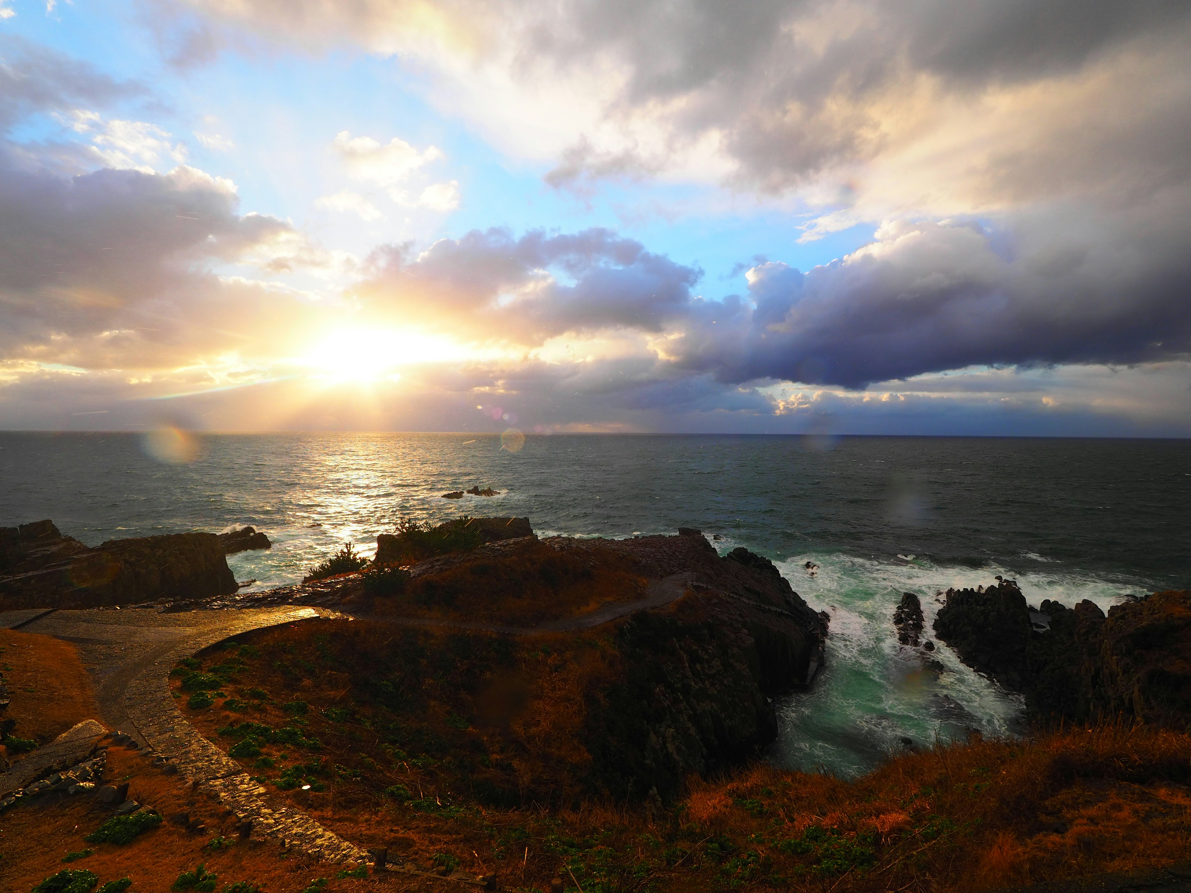 海岸風景，夕陽穿透雲層，波浪拍打岩石