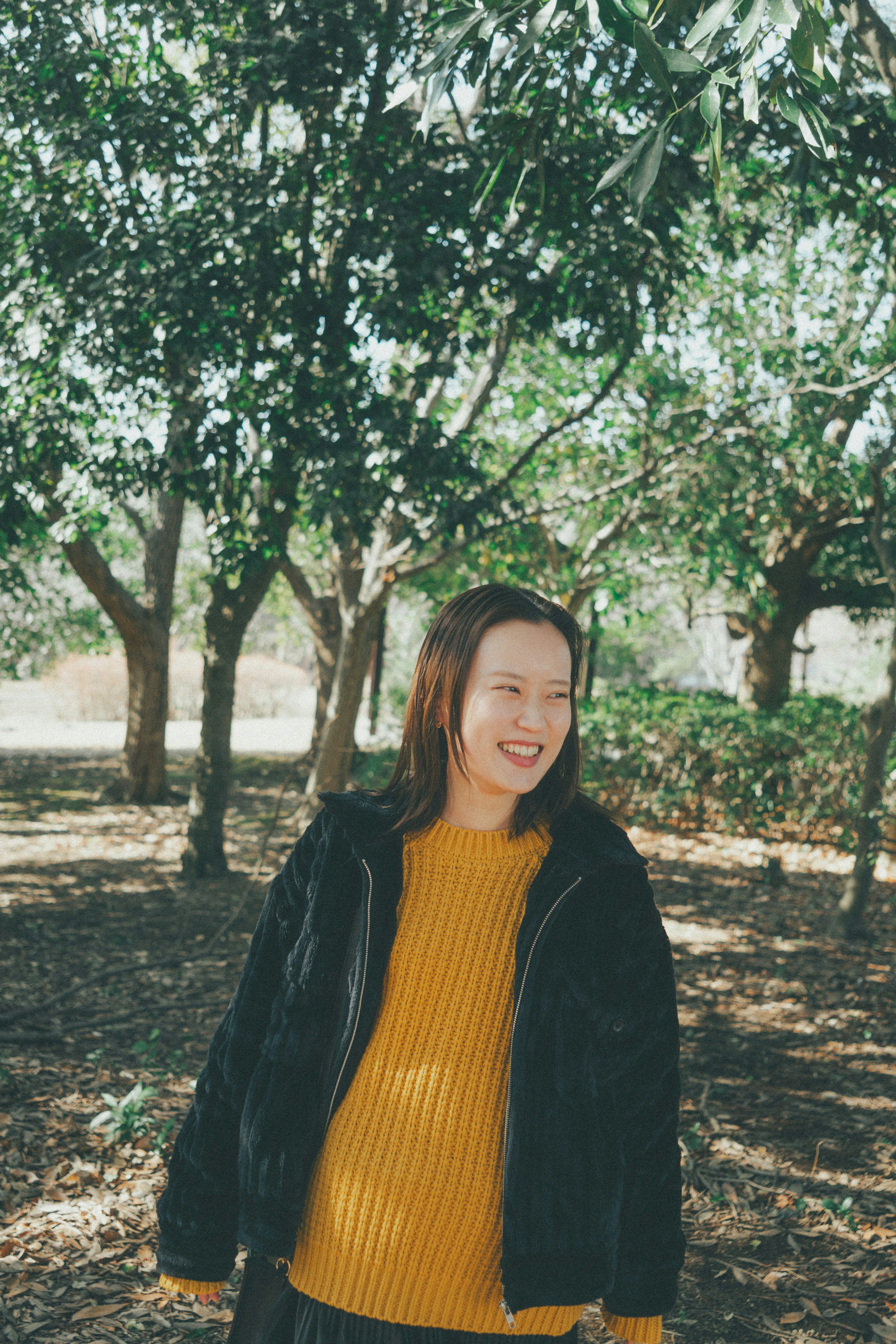 Une femme souriante se tenant parmi des arbres, portant un pull jaune et une veste noire