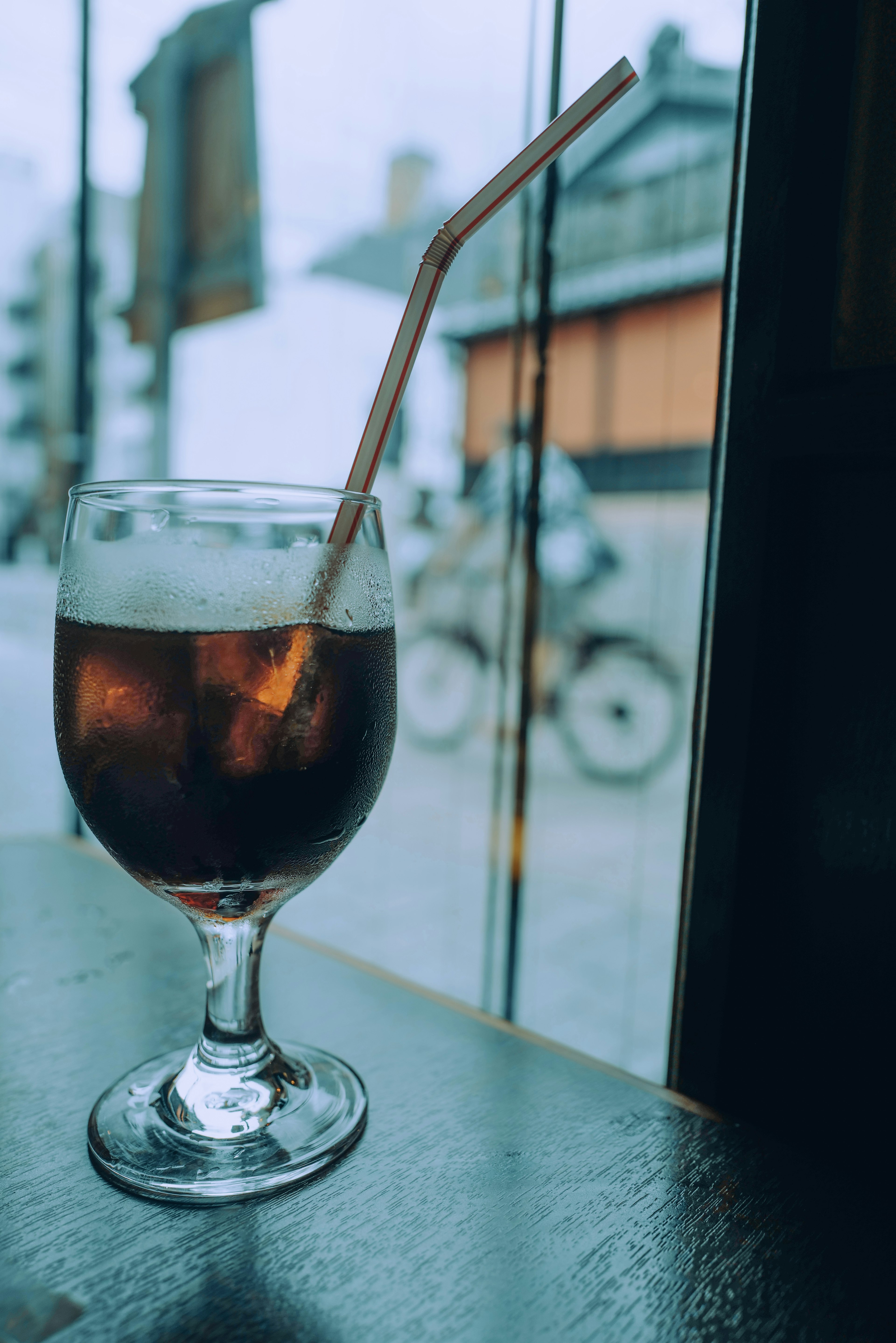 Un vaso de cola con una pajilla en una mesa cerca de la ventana