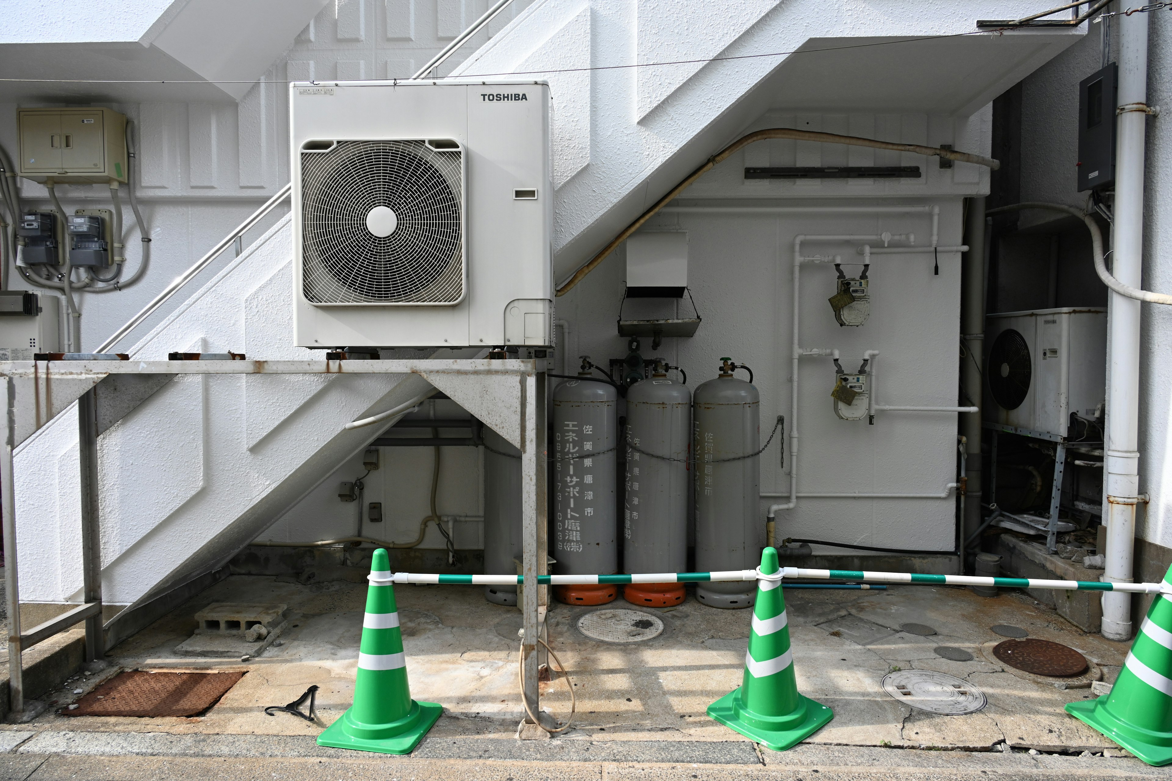 View of an air conditioning unit and gray cylinders under a staircase green cones and white wall