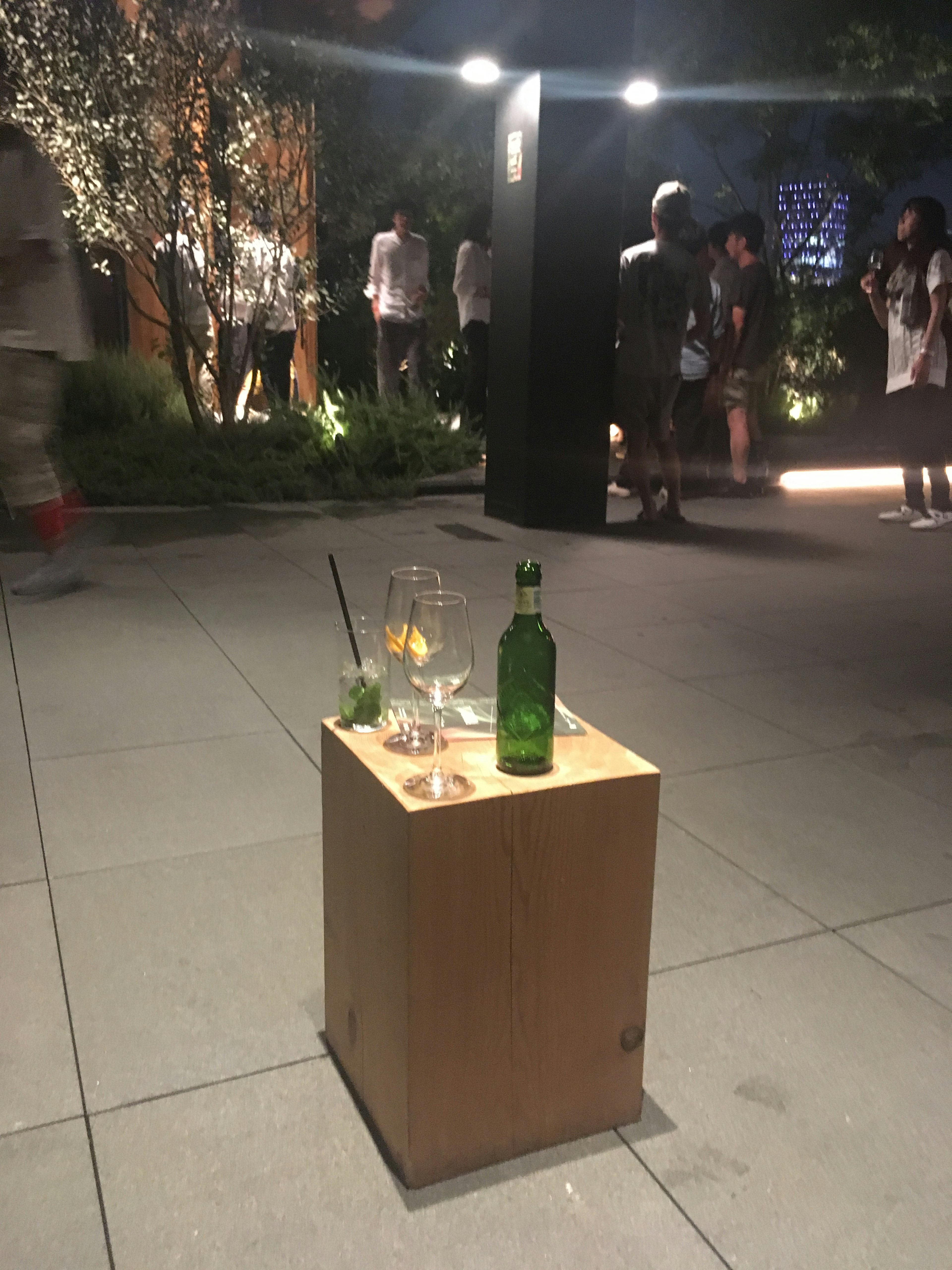 Pedestal de madera con un vaso y una botella verde en un evento nocturno al aire libre