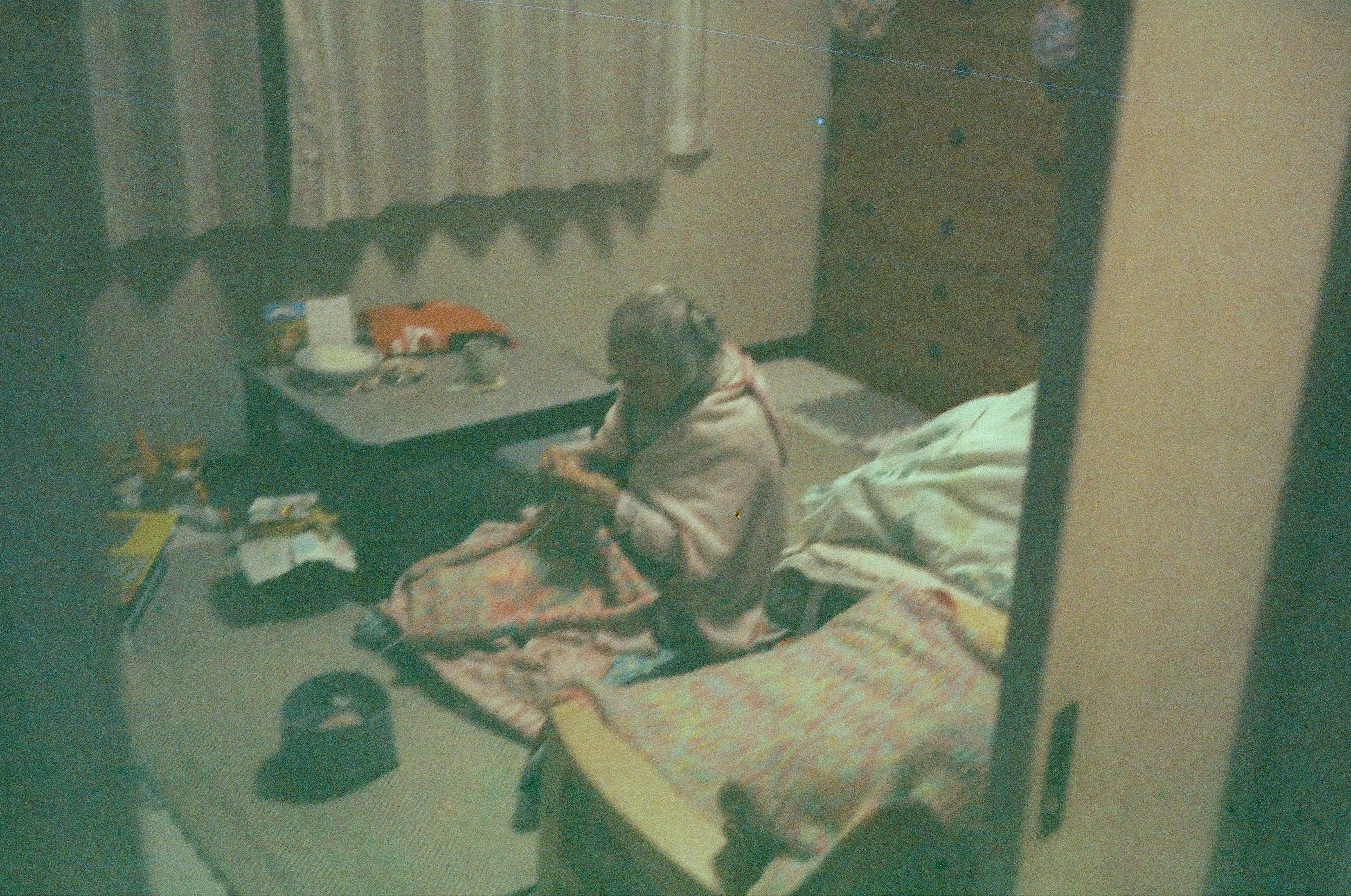 An elderly person sitting on a bed wrapped in a blanket in a dimly lit room