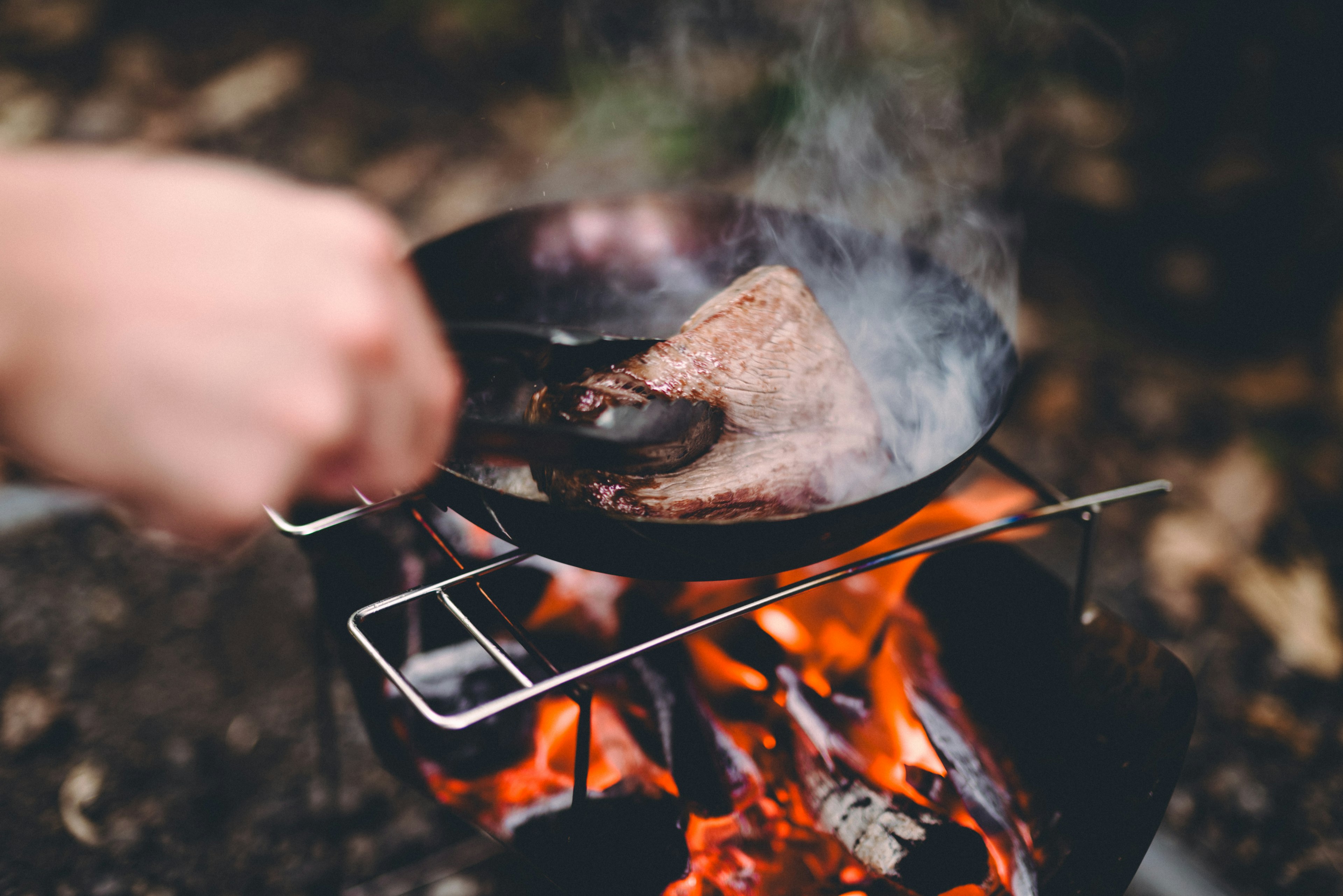 Mano sosteniendo una sartén con carne cocinándose sobre una llama abierta