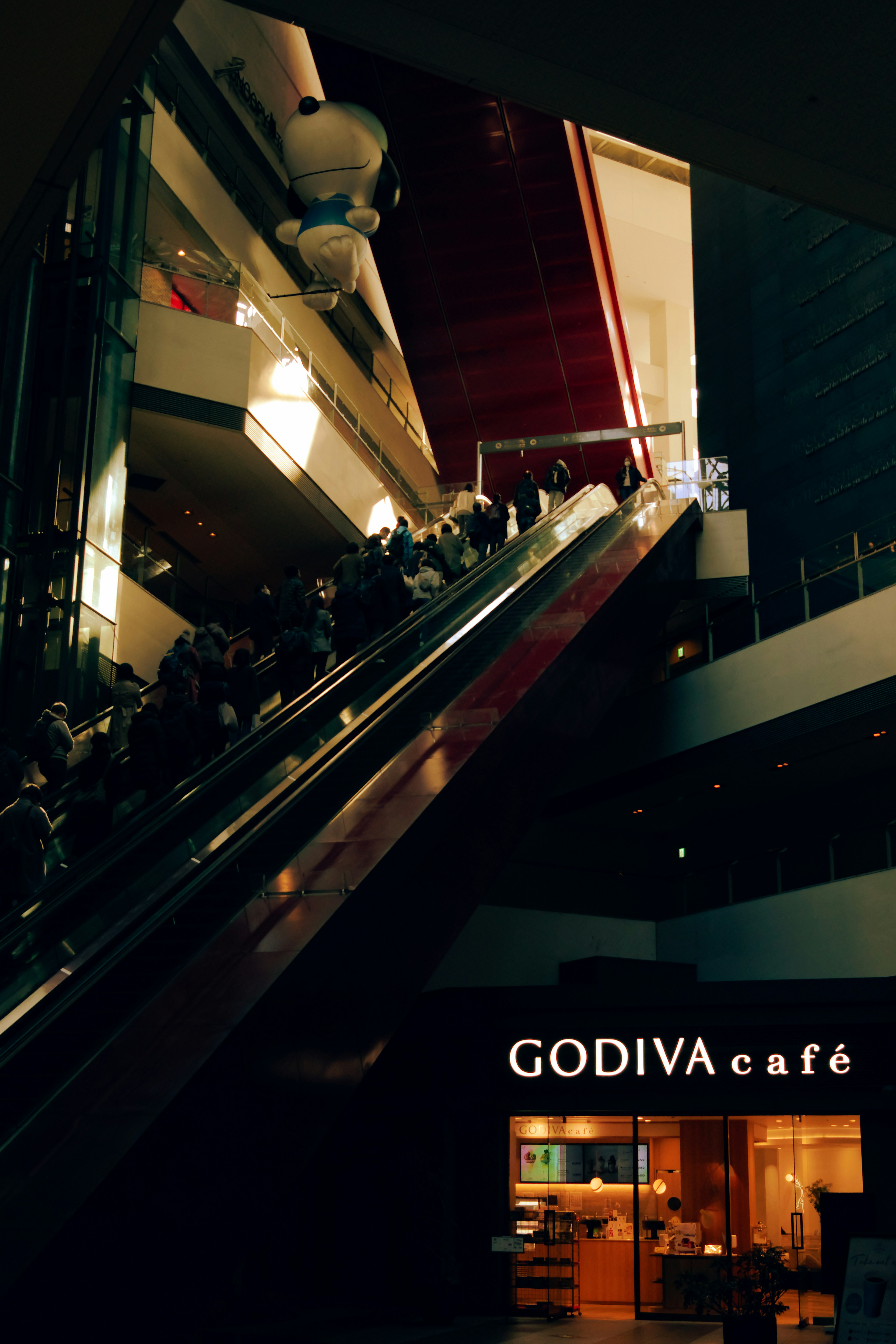 Interior of a shopping mall with people on an escalator and a GODIVA café