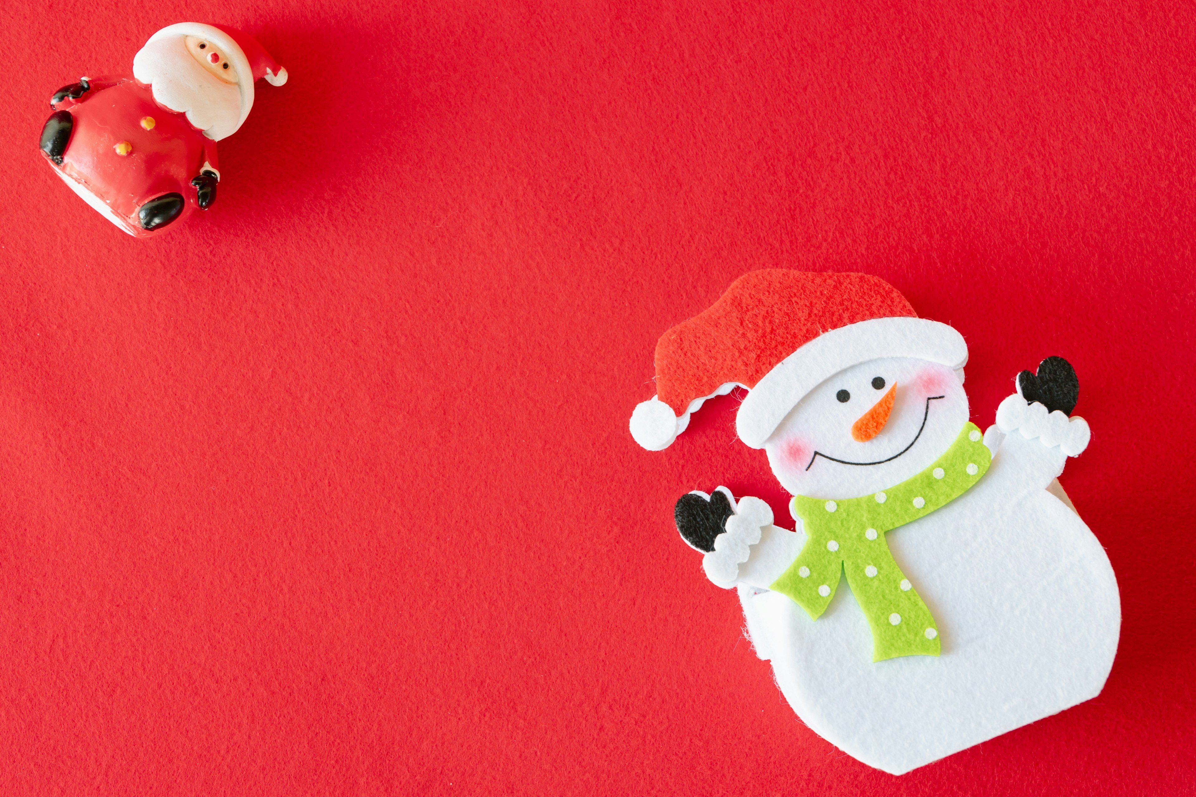 A cheerful snowman with a Santa hat and a green scarf on a red background alongside a small Santa decoration