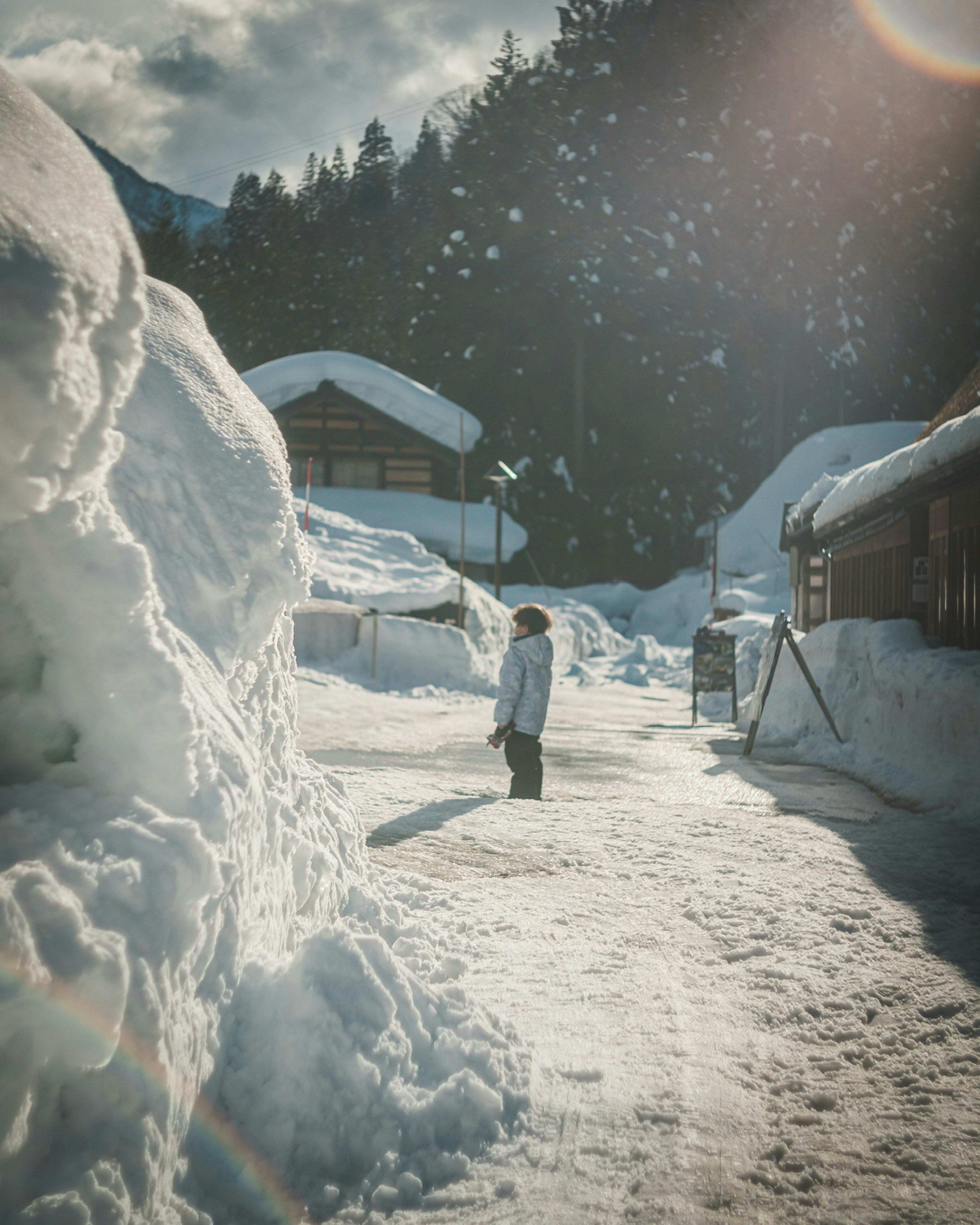 雪に覆われた村の通りで立つ人物　周囲には雪山と木製の家々