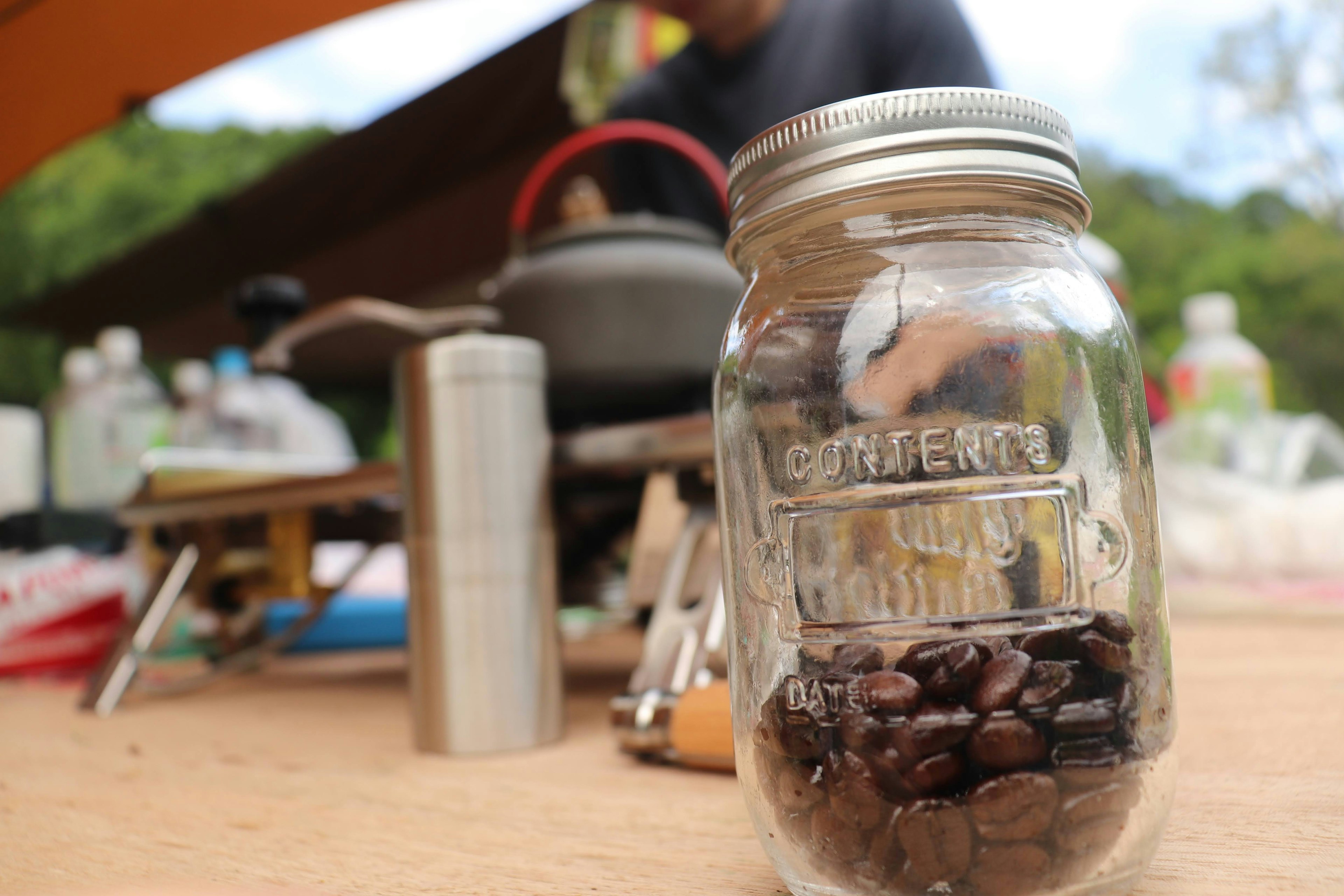 A glass jar filled with coffee beans placed on a table with outdoor cooking equipment in the background