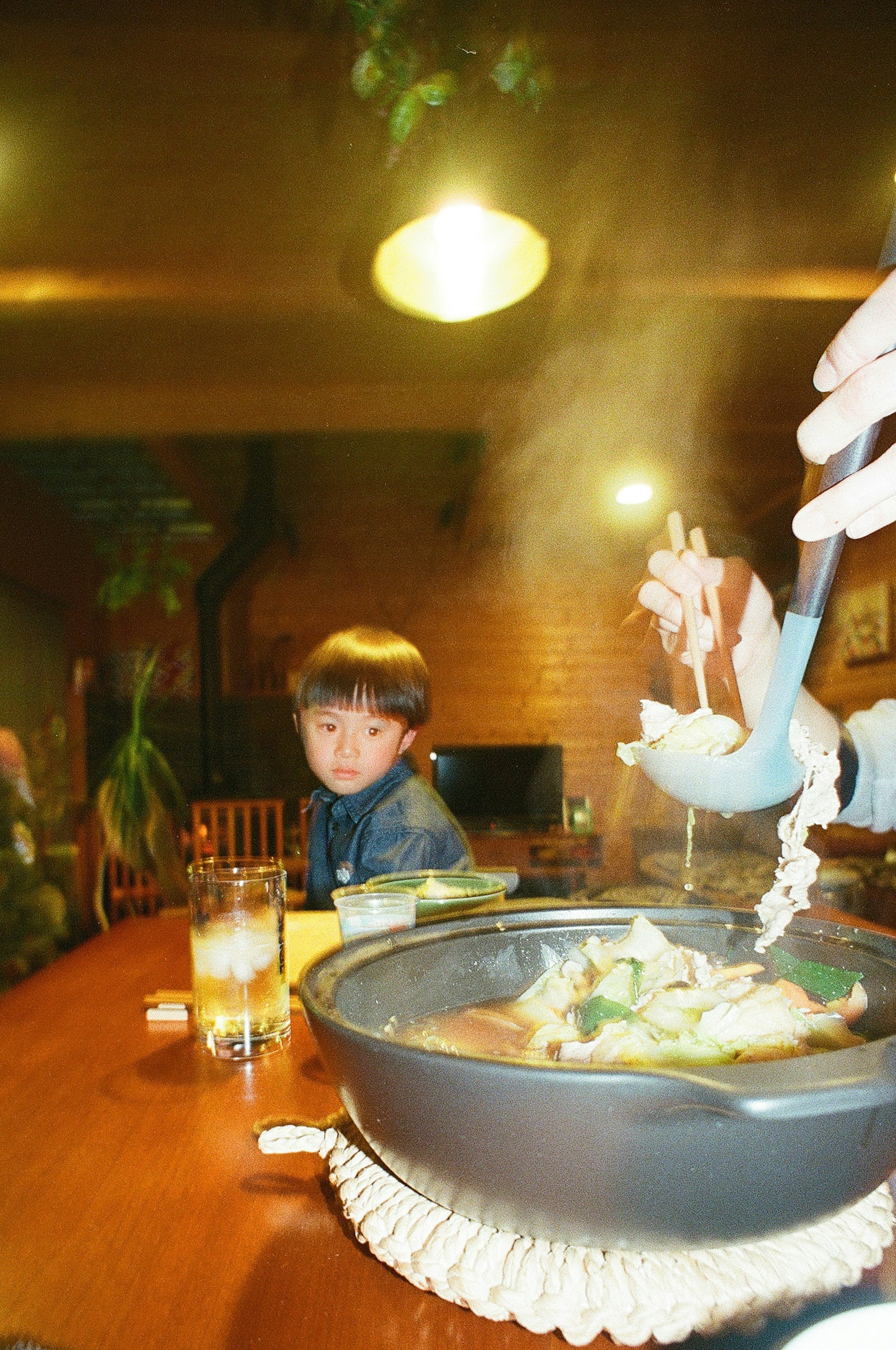 A scene with a child sitting at a table and a hot pot dish steaming with ingredients being served