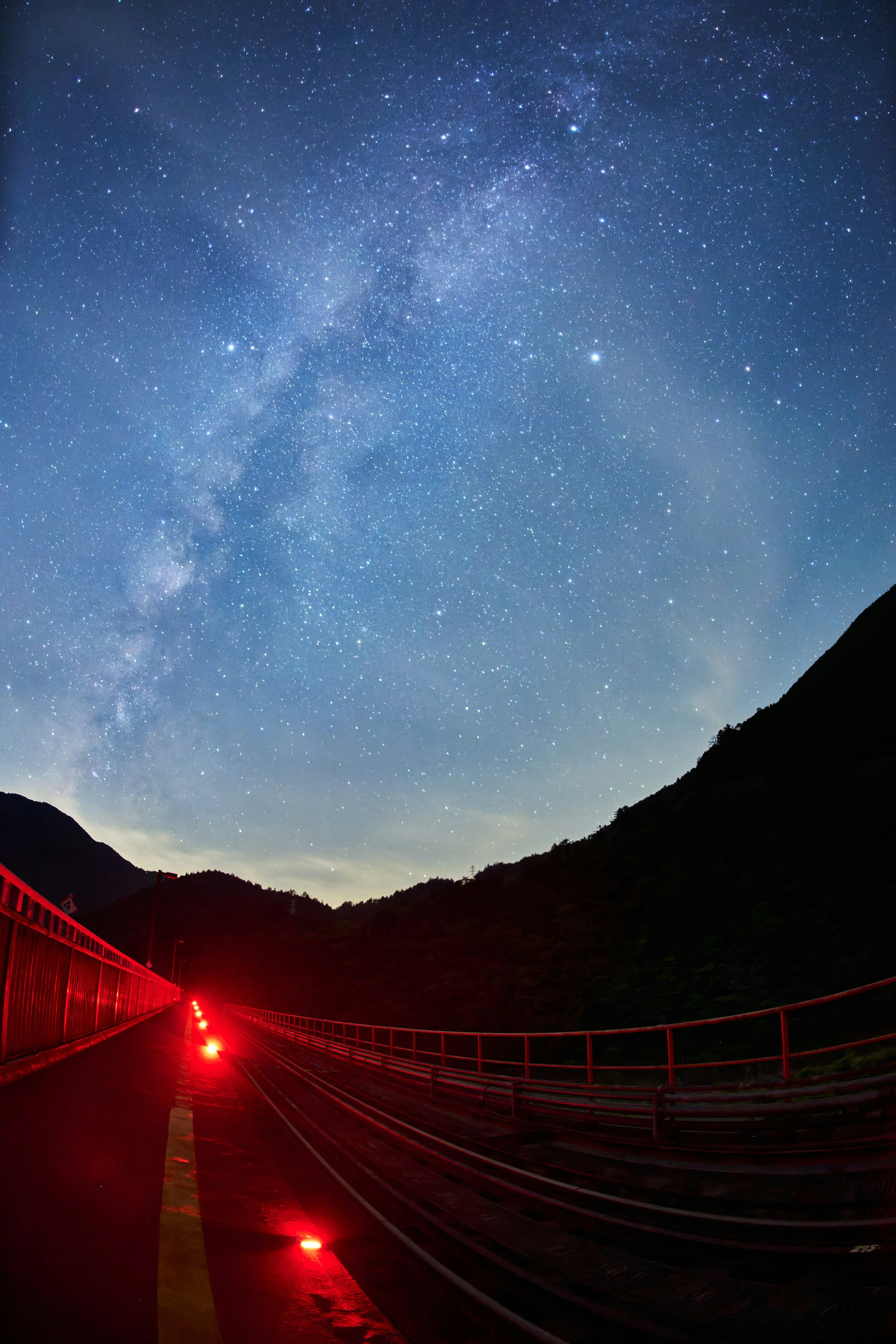 Camino de montaña con luces rojas bajo un cielo estrellado con la Vía Láctea