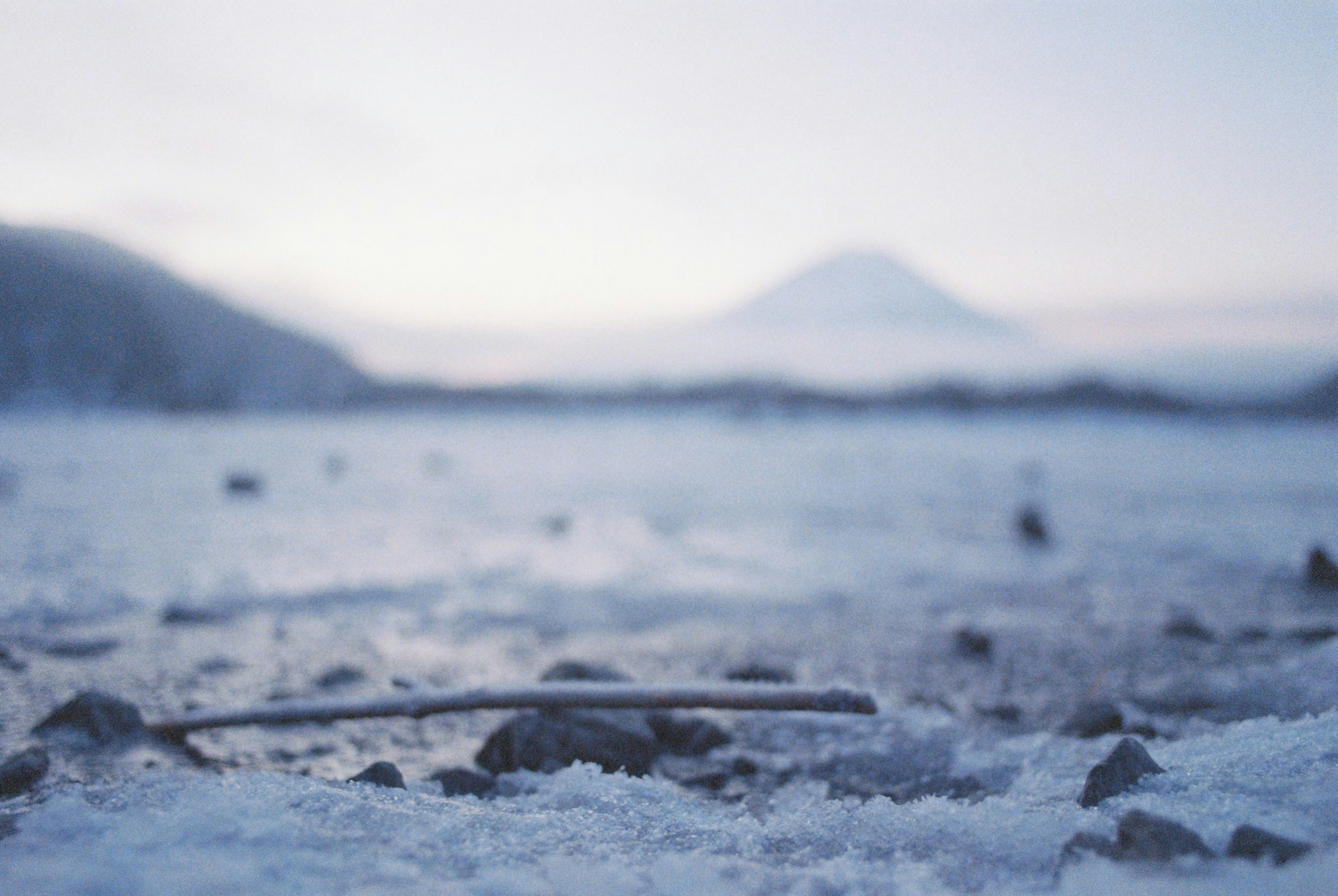 Gros plan de galets et d'une brindille sur une rive enneigée d'un lac avec le mont Fuji en arrière-plan dans un paysage d'hiver