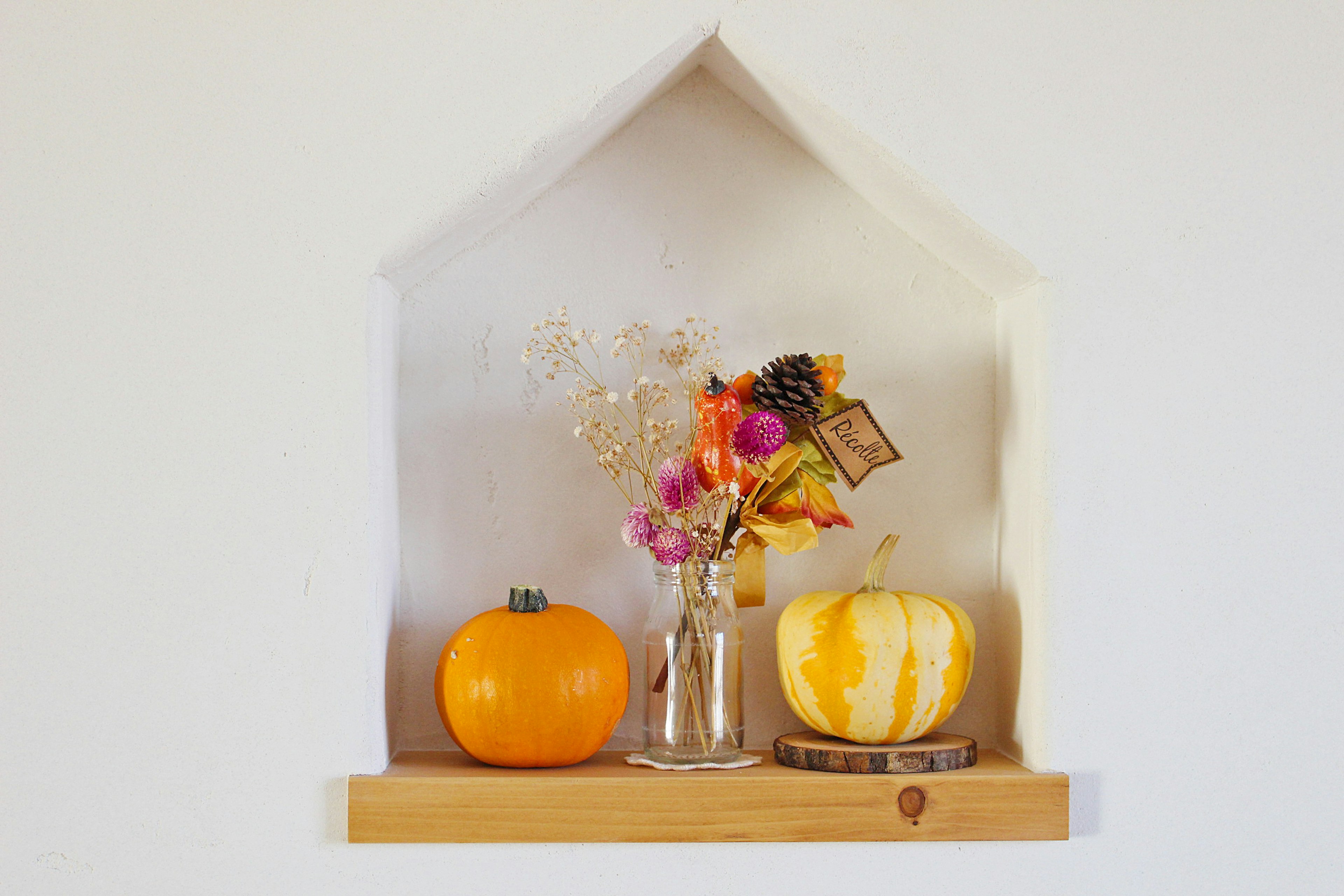 Étagère décorative sur un mur blanc avec des décorations d'Halloween comprenant des citrouilles orange et un vase de fleurs séchées