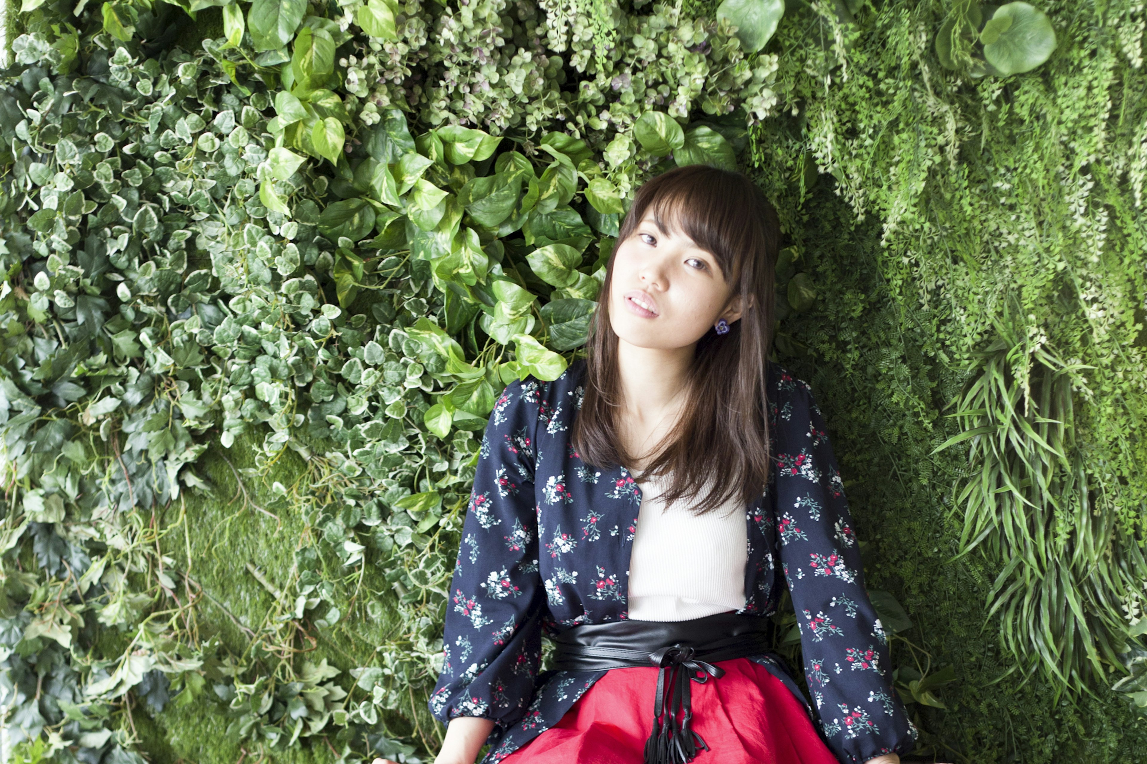 A woman sitting in front of a green wall wearing a floral jacket and red skirt