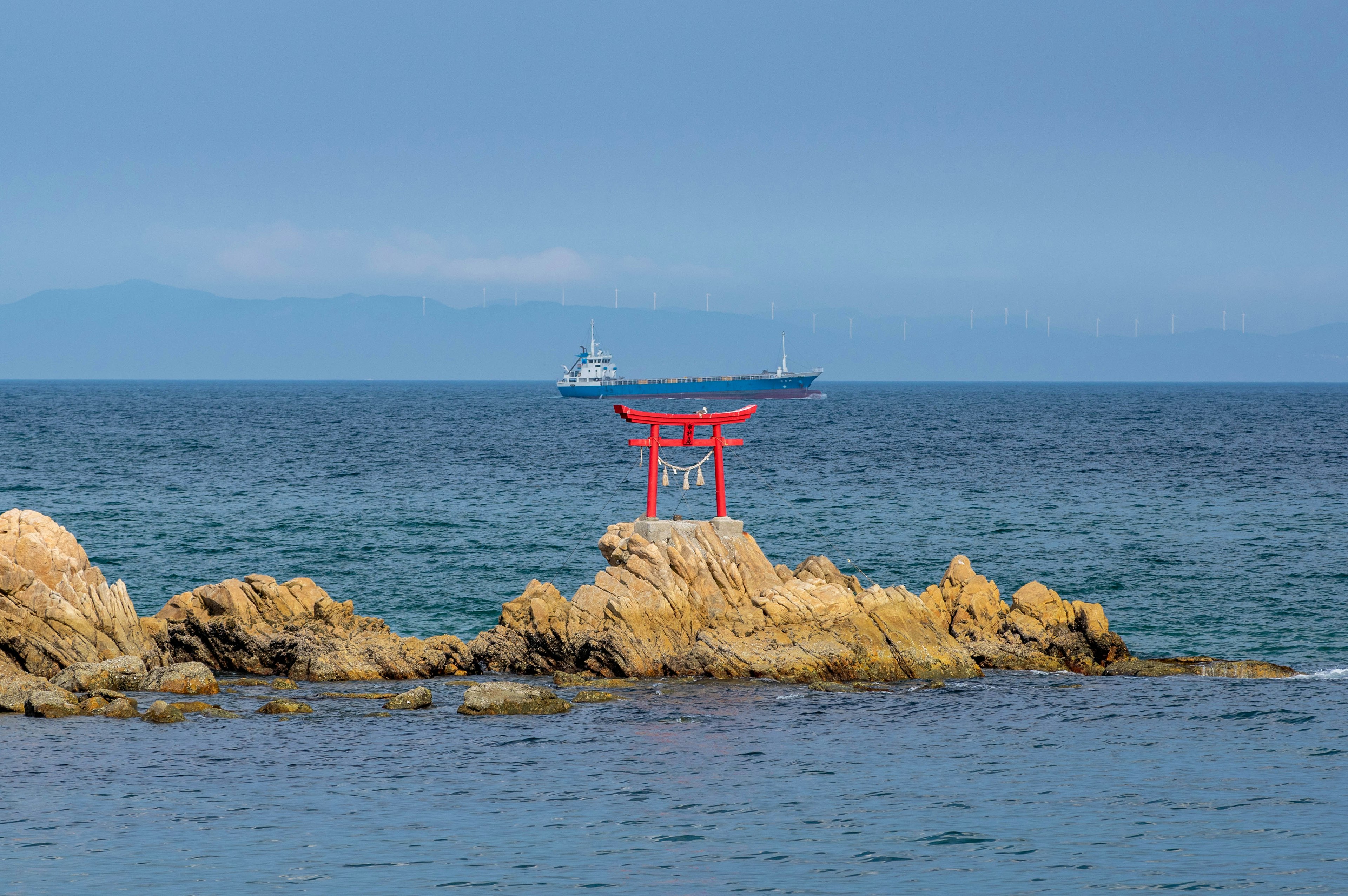 Un torii rosso si erge su rocce nell'oceano con una nave sullo sfondo